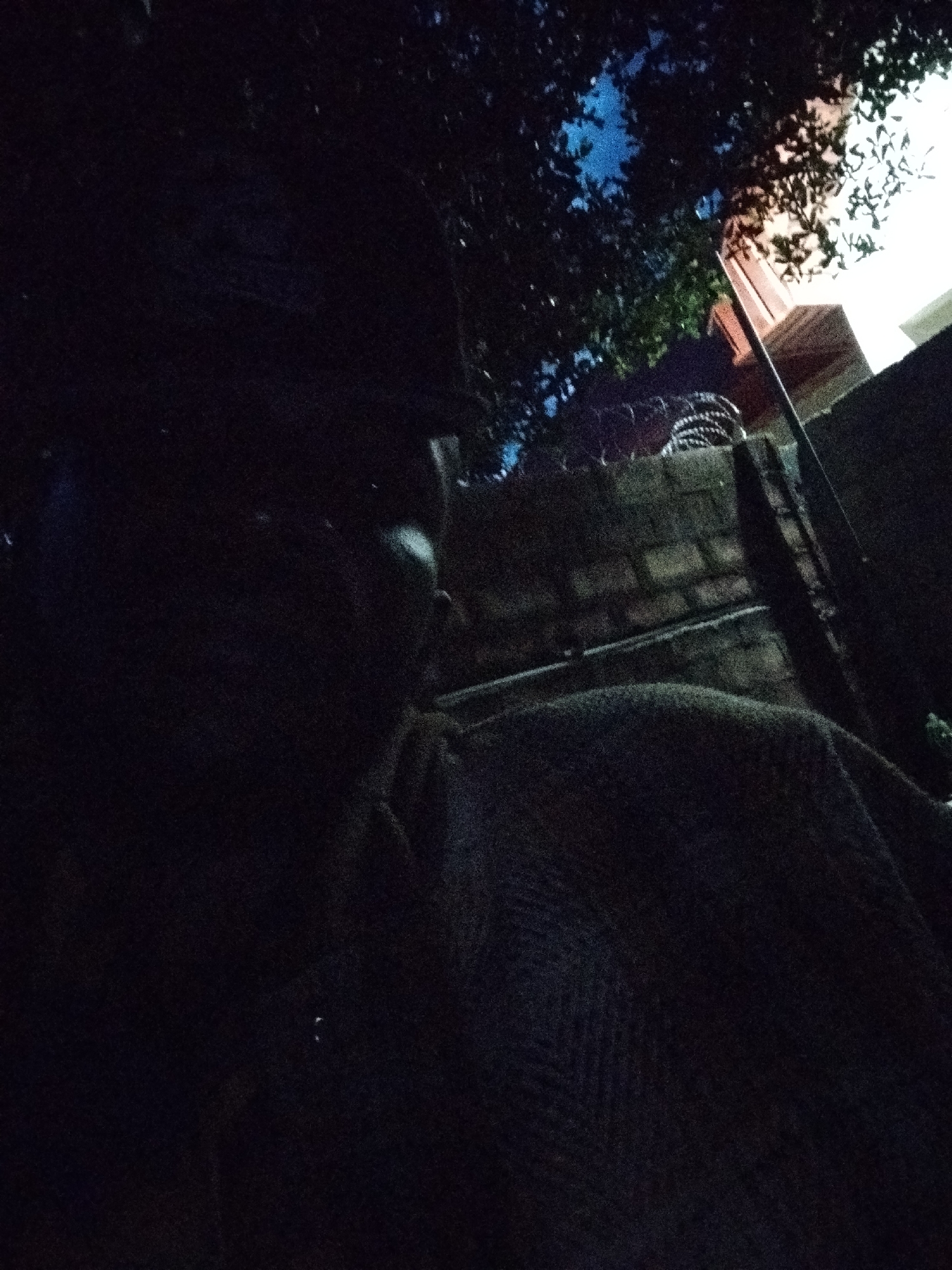 The image depicts a dark, low-light scene, likely taken at night or in a dimly lit indoor space. The primary subject appears to be a person, or at least part of a person's head and shoulder, visible on the left side of the frame. They are largely obscured by shadow, making specific details difficult to discern. A textured fabric, possibly a blanket or clothing, covers a significant portion of the lower right side of the image, suggesting the person is draped in it or resting.
In the background, there are hints of an outdoor setting. A brick wall is visible, with what appears to be barbed wire or a similar protective fencing material stretched across its top. Above the wall, foliage from trees is silhouetted against a bright, possibly artificial light source, which also illuminates a portion of a building with a reddish-brown facade. The sky above the trees shows a deep blue, suggesting it is nighttime.
The overall mood is somewhat mysterious and intimate, due to the darkness and the close-up perspective. There is no visible text in the image. The location context of Mangu Town, Nigeria, is noted but not visually evident from the image itself. The activity is unclear, but it seems to be a moment of rest or quiet observation.