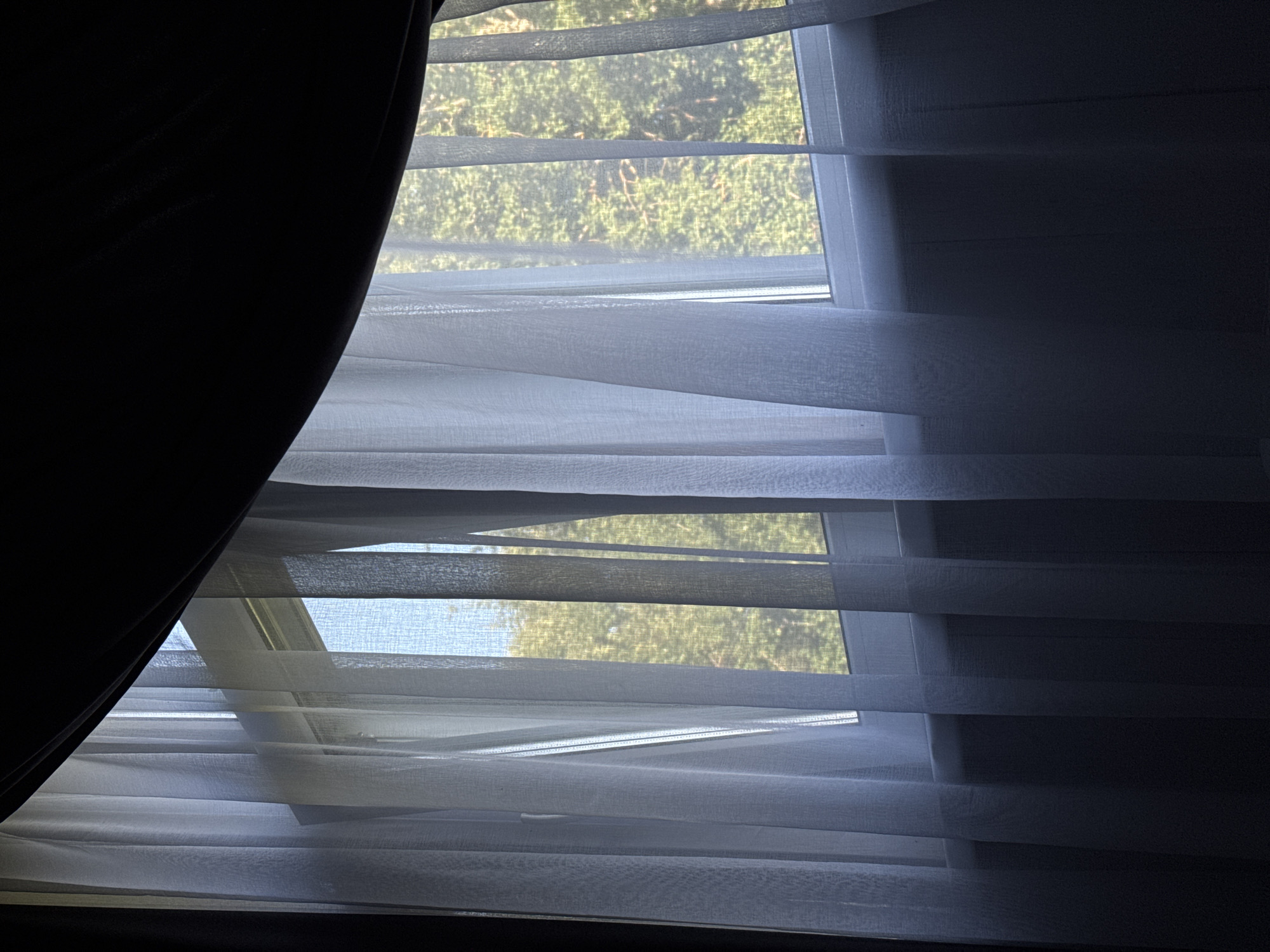 A view through a sheer white curtain reveals a window with trees and sky visible beyond. The curtain is partially drawn, creating vertical folds that obscure parts of the view. A dark curtain is visible at the top of the frame, suggesting it is partially closed, or a separate layer of window covering. The overall impression is one of a domestic interior looking out onto a natural scene. The location in the city of Doma 1096 Km, Russia is not discernible from the image.