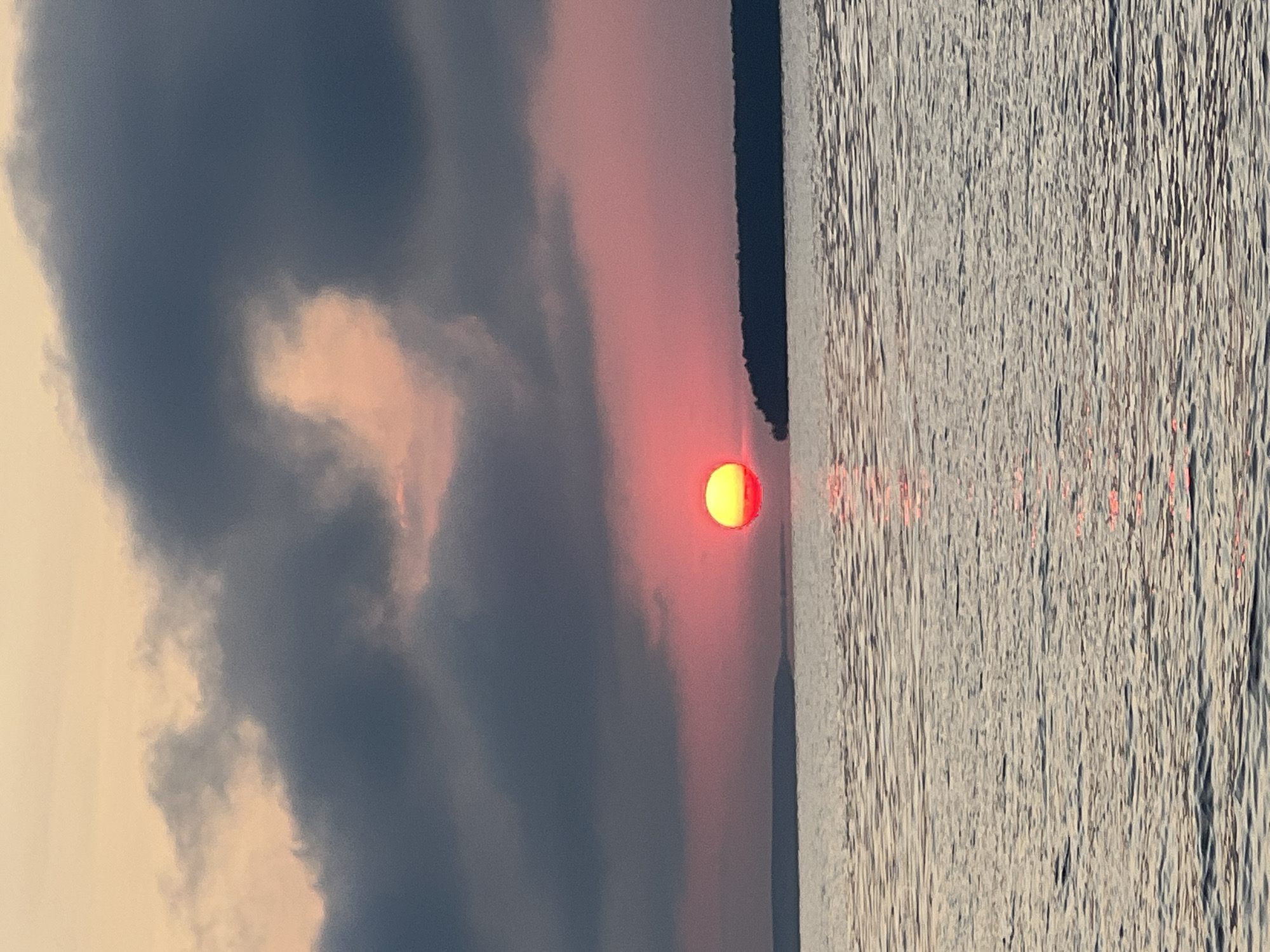 A sunset is visible over a body of water, with a sliver of land on the horizon. Dark clouds are present in the sky, obscuring some of the light from the setting sun. The water is calm and reflects the colors of the sunset. The scene is one of natural beauty, with the sunset creating a dramatic and colorful display. The location is the village of Novopichugovo, Russia.