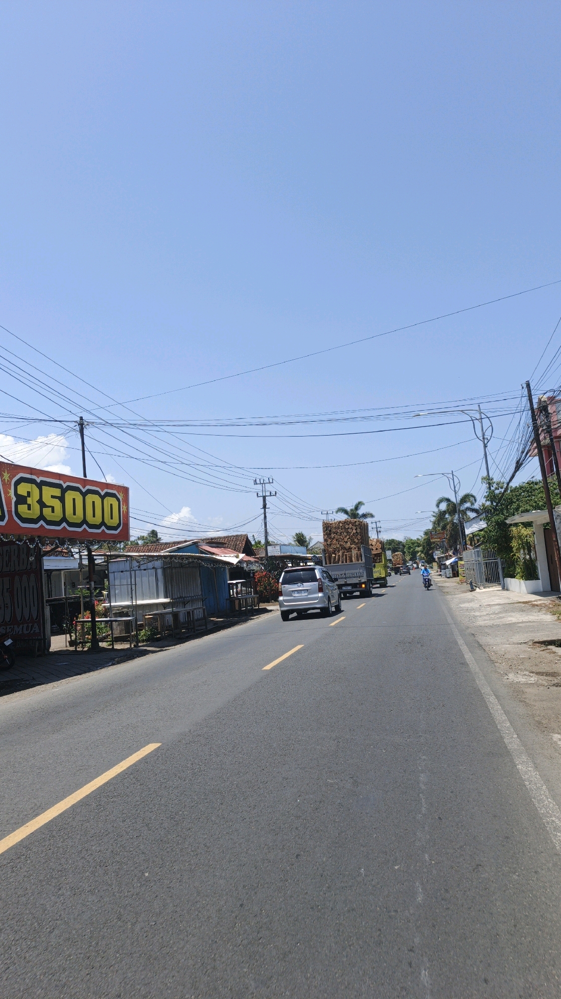 The image depicts a street scene in Cikembulan, Indonesia, on a clear, sunny day. The road, paved with asphalt, has yellow lane markings. On the left side of the road, there are several small roadside stalls with simple structures and a large yellow sign advertising "35000". A silver minivan is visible traveling on the road, followed by a truck carrying a large load of logs. Further down the road, a person on a motorcycle is also seen. The environment includes scattered trees and vegetation, utility poles with numerous overhead wires, and buildings lining the street, suggesting a semi-urban or rural commercial area.
