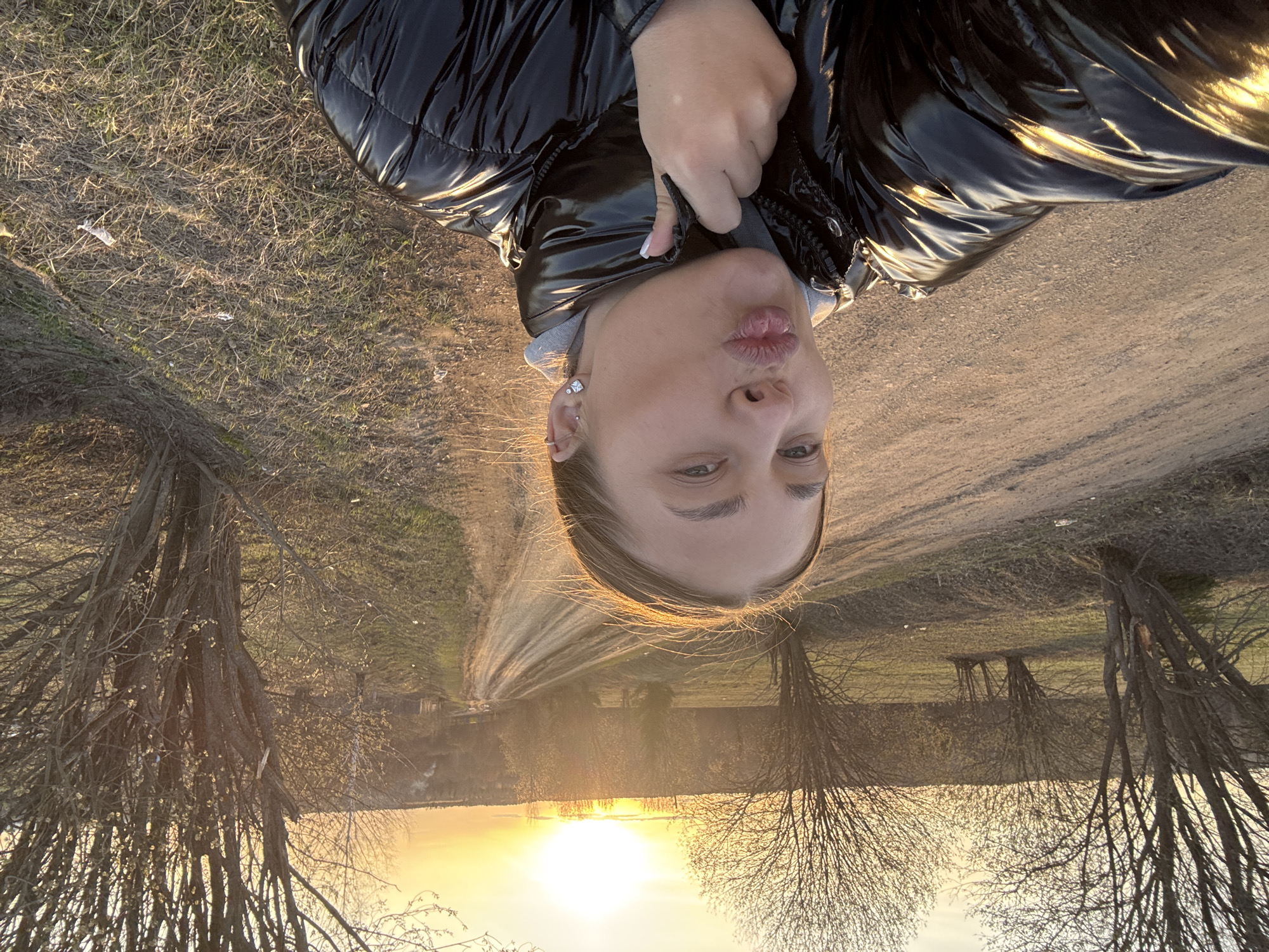 The image shows a woman in a black puffer jacket taking a selfie. She is facing the camera with a slightly puckered expression. The background depicts a dirt road leading towards a setting sun reflected in a body of water. Bare trees line the road. The location is Amin'kai, Russia.