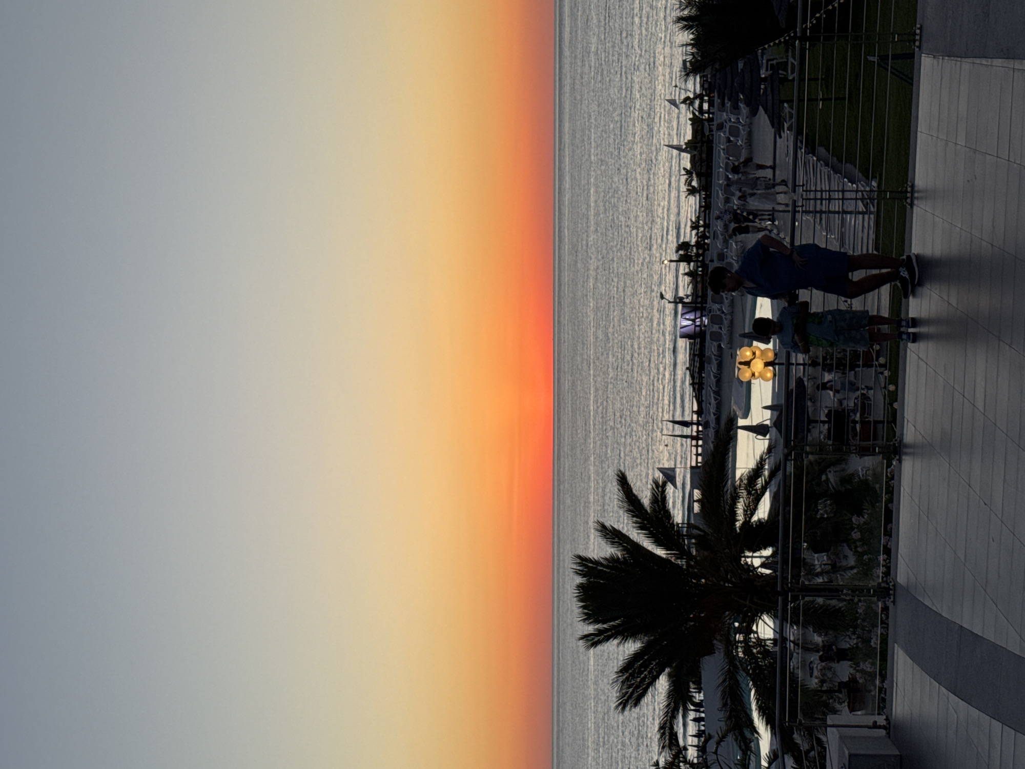 This image captures a serene sunset scene overlooking the Mediterranean Sea from a vantage point in Isola delle Femmine, Italy. The sky is a gradient of soft blues and oranges, transitioning into a fiery red band just above the horizon. The sea is calm, reflecting the colors of the sunset.
In the foreground, a silhouetted palm tree stands prominently on the left side of the frame. Beyond it, a resort area is visible with lounge chairs and umbrellas, suggesting a vacation destination. Two figures are silhouetted on a balcony in the middle ground, one an adult and the other a child, appearing to observe the sunset. Their presence adds a human element to the tranquil landscape.
The overall atmosphere is peaceful and picturesque, embodying the beauty of a Mediterranean sunset.