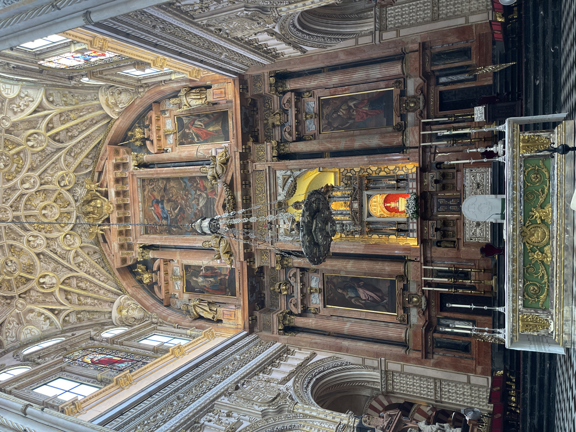 The image depicts the interior of the Mezquita-Cathedral of Córdoba, Spain, specifically focusing on the altar and its surrounding area. The central focus is a grand altar with a marble top and a green, gold-adorned front. Behind the altar, a richly decorated reredos features multiple paintings and sculptures. A large, ornate chandelier hangs from the intricately decorated ceiling, which is adorned with gold accents and religious motifs. The walls are lined with marble and feature stained-glass windows. In the foreground, a checkered floor is visible. No people are present in the frame. The scene conveys a sense of historical and religious grandeur, typical of a cathedral setting.