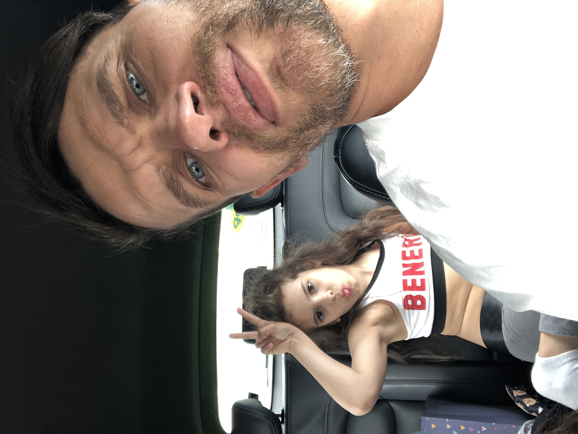 This media file depicts a selfie taken inside a vehicle, with the camera oriented vertically and the subjects appearing upside down. The foreground features the upper body and face of a man with light skin, dark hair, and blue eyes. He has stubble on his chin and mouth area and is wearing a white t-shirt. His expression appears playful. In the background, a young girl with long, curly brown hair is lying down on the car seat. She has fair skin and brown eyes, and is making a peace sign with her left hand while sticking out her tongue. She is wearing a white sports bra with red lettering that reads "BENER" and grey leggings with white socks. The interior of the car is visible, with dark leather seats and a white background that suggests a car window. The scene appears to be an indoor setting within a vehicle, possibly during the daytime due to the lighting. No specific weather cues are apparent. The overall emotion conveyed is one of casual fun and a shared moment between the two individuals.