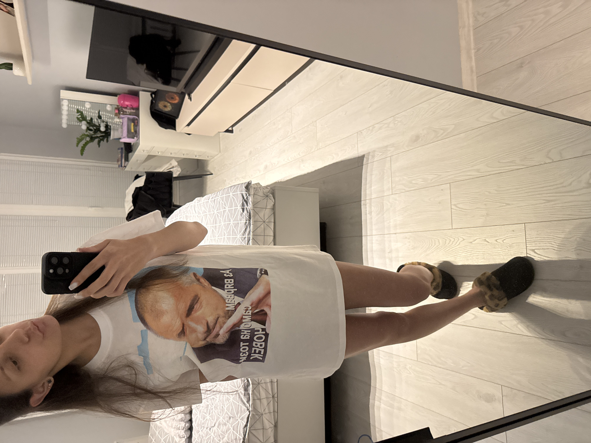 A person stands in a room, reflected in a mirror. They wear a white t-shirt with an image of a man, dark-colored slippers, and hold a smartphone in their left hand. The room includes a bed with a gray and white geometric patterned duvet cover, light-colored hardwood flooring, a white cabinet with a television on top, and a small vanity with decorative items. The location is likely a residential interior, although Sofia, Bulgaria cannot be identified.