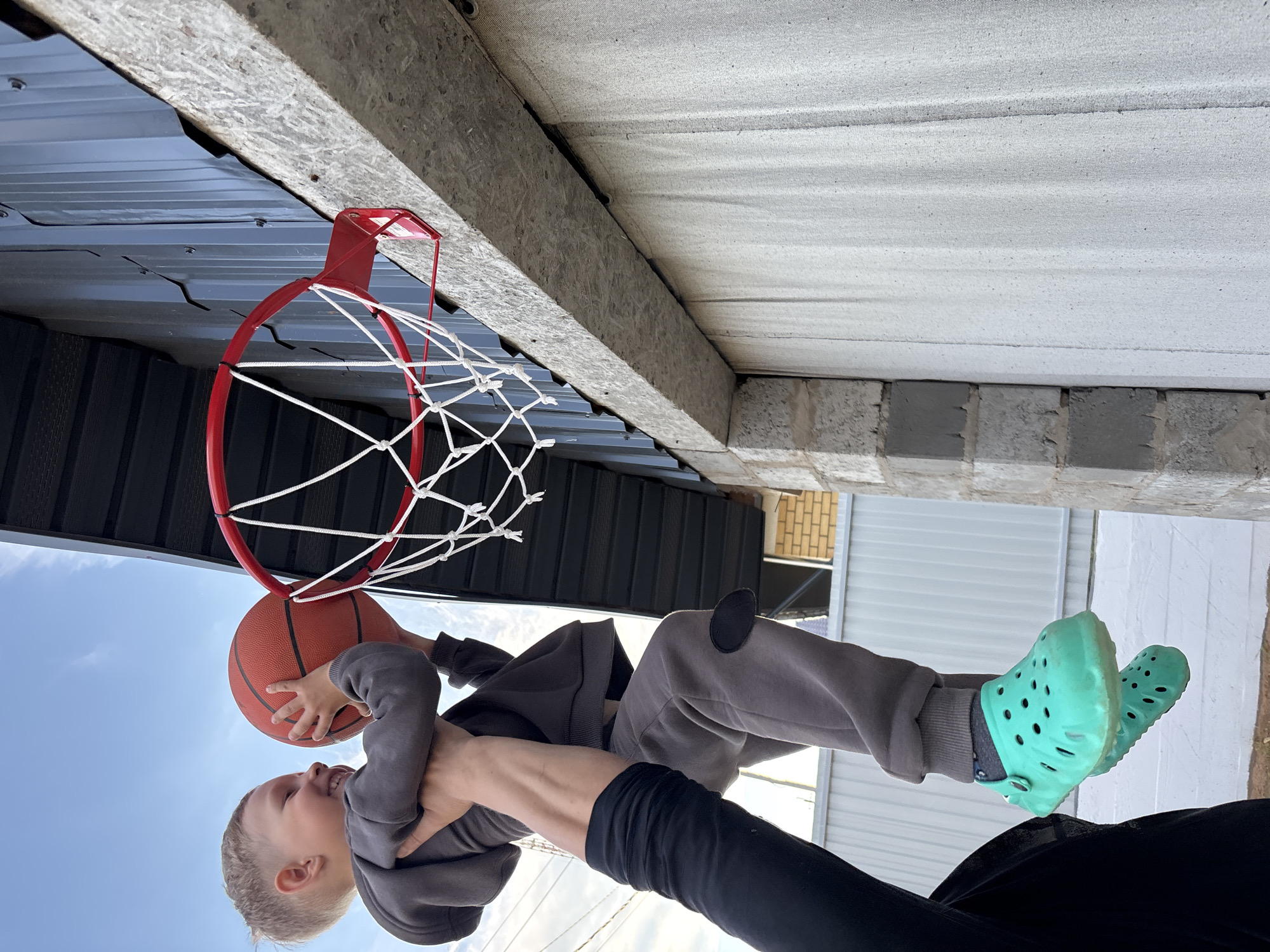 An outdoor scene, likely captured in Chaikovsky, Russia, shows a playful interaction between an adult and a young child involving a basketball hoop. An adult, visible only by their arms and upper torso in black long-sleeved clothing, lifts a young child towards a red-rimmed basketball hoop with a white net.
The child, with light-colored hair, is smiling broadly and holding an orange basketball with both hands, positioned as if about to dunk or having just dunked it. The child wears a grey hooded sweatshirt, grey sweatpants, and distinctive turquoise or mint green Crocs.
The basketball hoop is mounted to a structure that includes a dark grey corrugated metal wall or roof section, beneath a thick, light-colored wooden beam (possibly OSB). Above the wooden beam, a textured, light-colored ceiling or wall surface, appearing to be made of concrete blocks or similar material, extends into the background.
A clear blue sky is visible, indicating a bright, sunny day. In the background, elements of other structures, such as white siding and a hint of brickwork, can be seen. A small white tag with red lettering is attached to the red bracket of the basketball hoop, but the text is not legible. The overall mood is joyful and active.