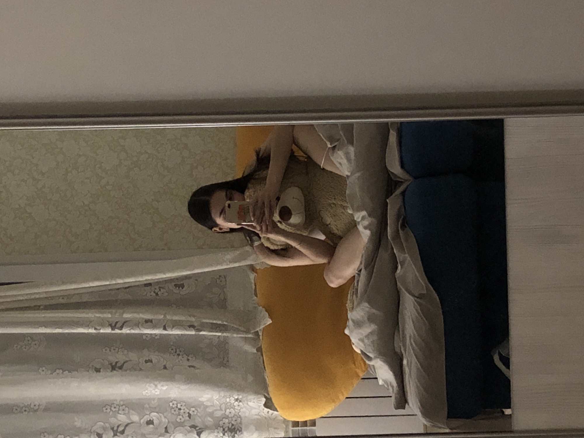 A reflection in a mirror shows a young woman lying in bed, possibly in Noviy, Russia. She is holding a phone in front of her face, likely taking a selfie. She is embracing a large teddy bear. The bed has a yellow pillow, a blue headboard, and a light-colored blanket. The wall behind the bed is decorated with floral wallpaper. Curtains are also present on the side.