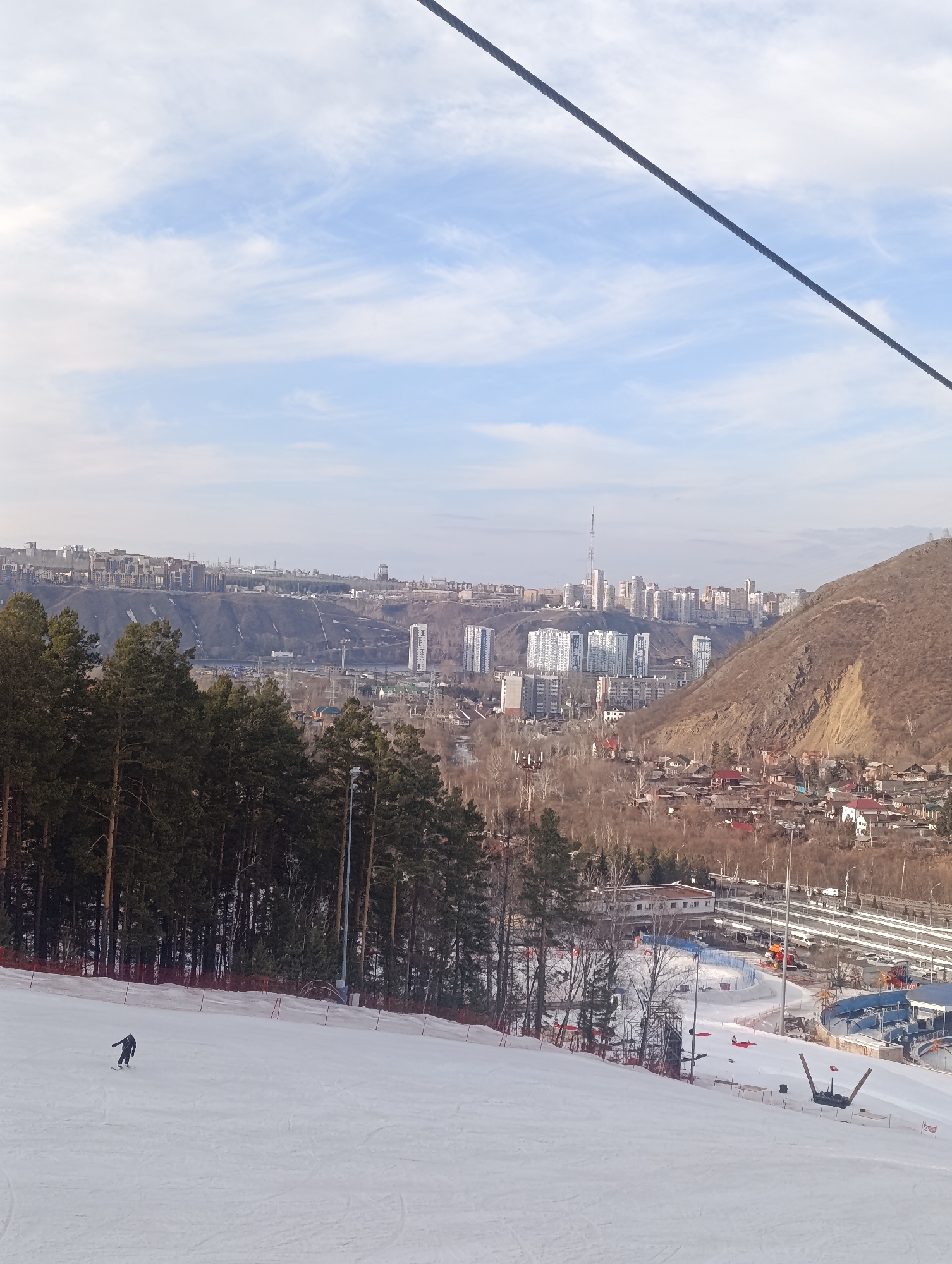 A wide, downward-angled view from a snow-covered ski slope in Krasnoyarsk, Russia, overlooks the city and a surrounding valley. In the foreground, a single individual in dark attire is snowboarding down the groomed slope, which is bordered by a red safety net on the right and dense pine trees on the left. The midground features ski resort infrastructure at the base of the slope, including structures, a visible ice rink with blue barriers, and assorted bare trees. To the far right, a steep, rocky, barren hillside rises abruptly. The background displays the extensive cityscape of Krasnoyarsk, marked by numerous multi-story residential and commercial buildings, a concentration of white high-rises, and a prominent communication tower. Distant hills and a partly cloudy sky form the horizon. A thick, dark cable diagonally crosses the upper right portion of the image.