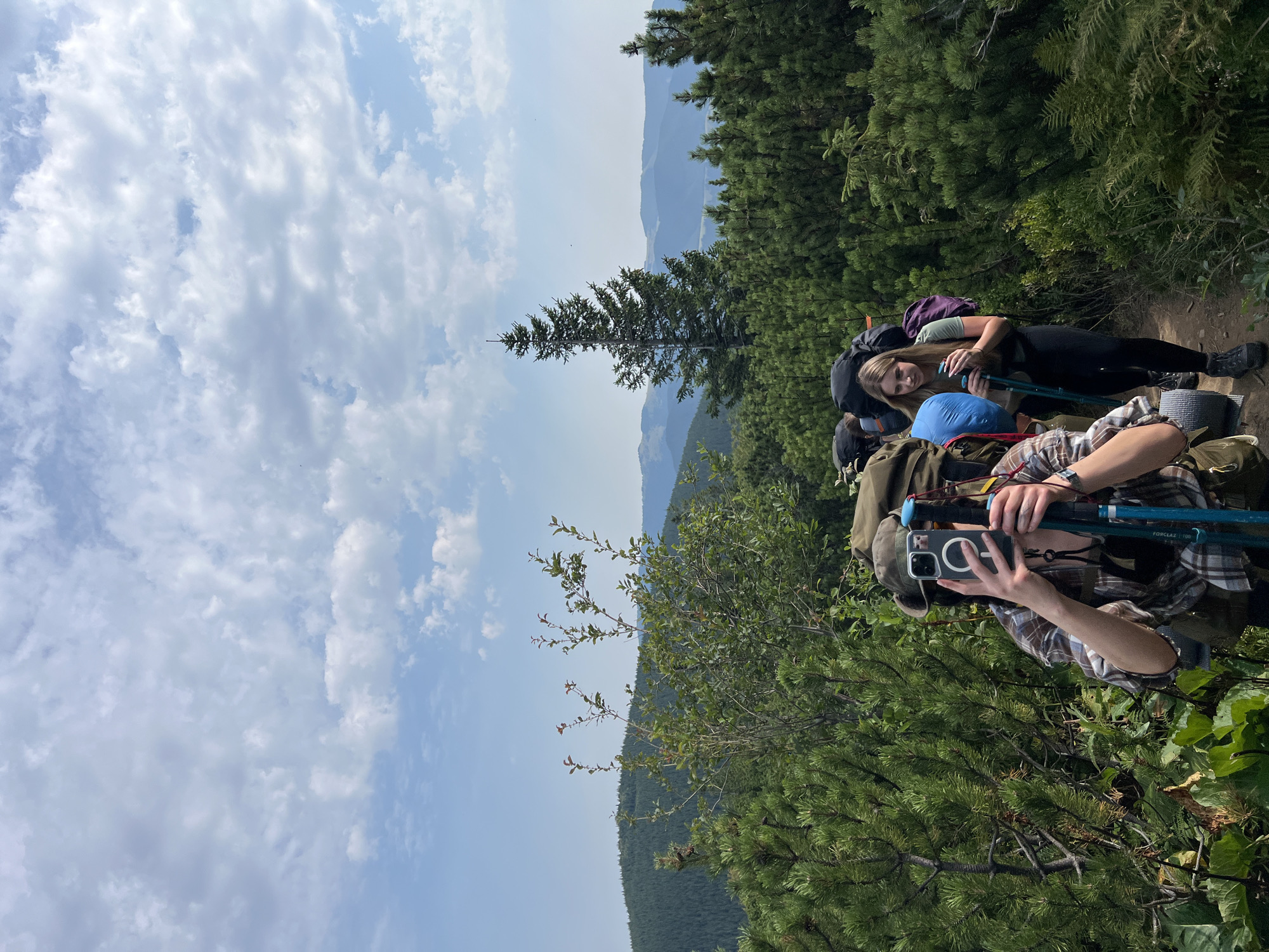 A group of hikers, two of whom are clearly visible, are ascending a mountain trail in a lush, green forest. The person in the foreground, wearing a plaid shirt and a hat, is holding up a smartphone to take a photo, while also holding trekking poles. Behind them, a young woman with blonde hair, dressed in a green t-shirt and black leggings, walks with trekking poles. Both individuals are equipped with large backpacks, indicating they are on a multi-day trek. The path is narrow and surrounded by dense pine trees and other vegetation. In the background, rolling hills covered in a dense forest stretch out towards the horizon under a partly cloudy sky. The scene appears to be located in the Carpathian Mountains, potentially near the town of Vorokhta, Ukraine, known for its scenic hiking trails. The time of day appears to be afternoon, judging by the lighting.