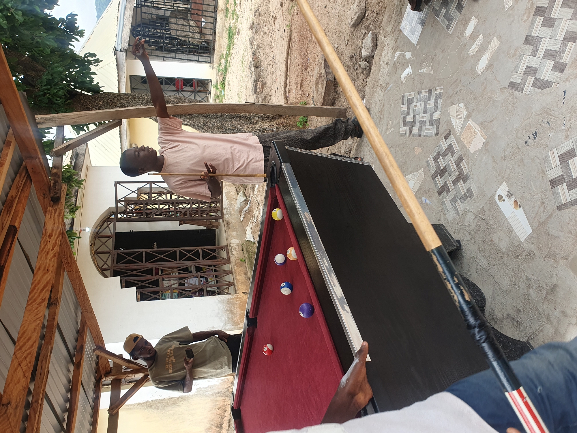 In Lokoja, Nigeria, under a rustic wooden and corrugated iron shelter, three young men are gathered around a red billiard table. One man, wearing a tan t-shirt and a baseball cap, stands to the left, looking down at his phone. To the right, another man in a pale pink t-shirt holds a pool cue, pointing with his other hand towards the background. A third person, partially visible on the left, appears to be assisting with the billiard table, with their hands near its edge. Several billiard balls are scattered across the table, including a white cue ball, a purple ball, a blue ball with the number 13, a red ball with the number 11, and an orange ball. The ground around the table is a mix of dirt and scattered floor tiles, suggesting an outdoor or partially constructed area. The background shows a building with a white facade and decorative metal gates, with trees and a distant hillside under a cloudy sky.