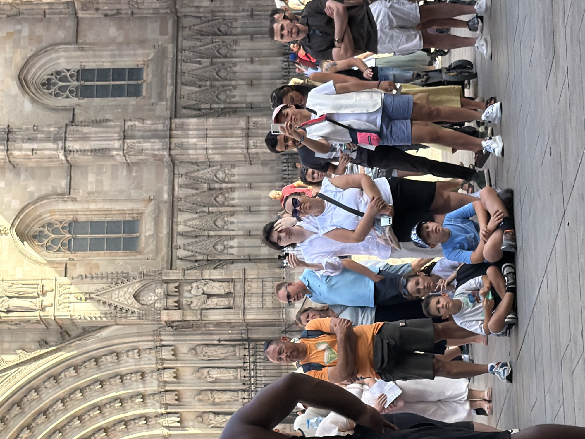 The image captures a crowd of tourists gathered on a paved plaza in front of Barcelona Cathedral. The cathedral's facade, featuring intricate Gothic architecture with arched doorways and statues, dominates the background. The foreground is occupied by a diverse group of people, including adults and children, some standing and some seated on the ground. Many appear to be observing something, with some holding phones and cameras. The atmosphere suggests a tourist attraction with a moderate level of foot traffic.