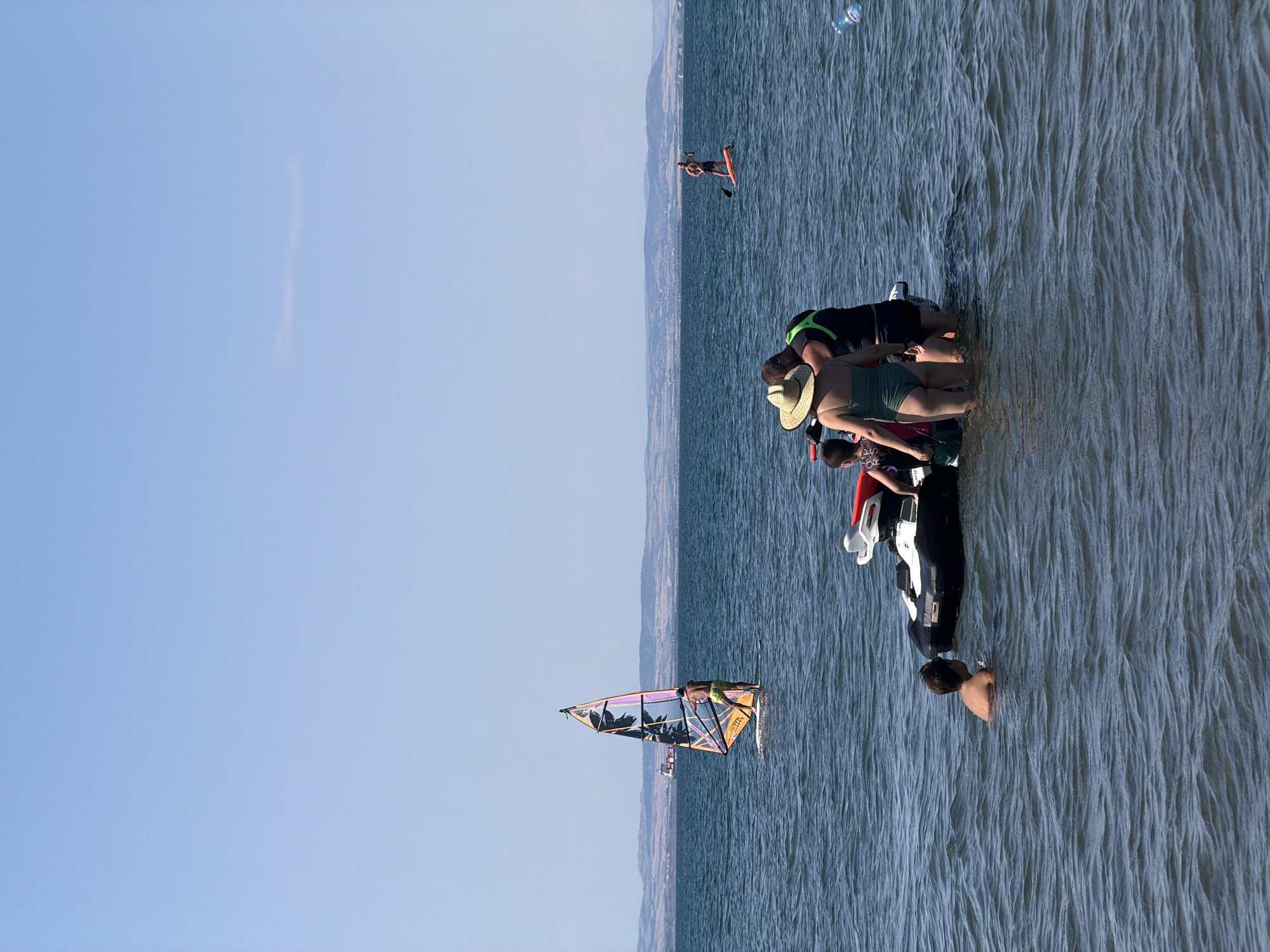 In Atiya, Bulgaria, a group of people are enjoying a sunny day at sea. In the foreground, a family stands in the shallow water next to a personal watercraft. A woman in a green swimsuit and a straw hat stands facing away from the camera, while a man in a life vest and shorts stands beside her, also facing away. A young boy in a dark swimsuit stands near them. In the middle ground, a windsurfer with a colorful sail navigates the waves, and in the distance, a person stands on a paddleboard. The sea is calm with gentle ripples, and the sky is a clear, bright blue. A cargo ship is visible on the horizon, near the coastline.