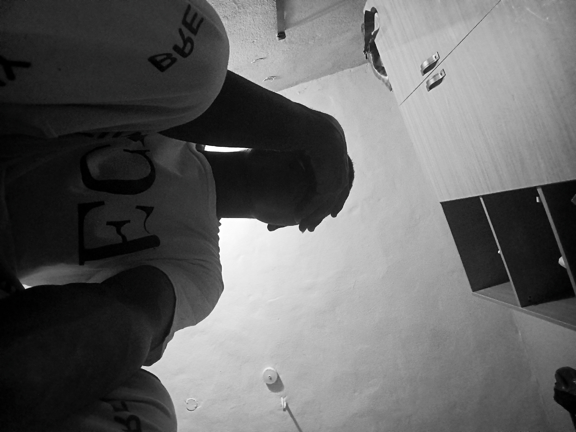 This black and white image is taken from a low, upward-looking angle, capturing an indoor scene.
A person is positioned on the left side of the frame, largely in silhouette against a lighter background. They are wearing a light-colored t-shirt and what appears to be light-colored shorts or trousers. Their head is bowed forward, with their hands clasped over it, suggesting a contemplative, distressed, or weary emotional state.
On the person's t-shirt, the letters "FC" are visible on the chest area. On their lower garment, the letters "BUE" are visible on the upper thigh. Both texts appear reversed from the viewer's perspective.
To the right, a light-colored wooden cabinet or wardrobe is visible, featuring two metallic handles. Below the cabinet, an open shelf unit or storage cubbies are present, rendered in darker tones. The background consists of a plain, textured wall. Near the bottom center of the frame, a circular light switch or similar wall fixture is mounted. A faint, small animal figure, possibly a cat, can be vaguely discerned near the top edge of the cabinet.
The monochromatic presentation contributes to a somber or introspective mood. The scene takes place indoors, likely within a room such as a bedroom or living space. There are no specific visual cues to indicate the time of day or weather.