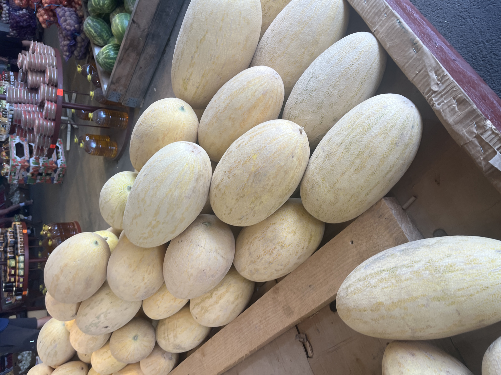 A market stall in Plastunovskaya, Russia, displays a large quantity of ripe cantaloupes. The cantaloupes are piled in wooden crates and on a wooden platform, with some of them partially obscuring each other. The fruits are oval-shaped with pale yellow skin and subtle vertical striping. In the background, shelves are stocked with jars of preserves and other produce, including watermelons and onions in netting. A person's legs are visible in the upper left corner, suggesting this is a functioning market. The lighting indicates it is daytime, likely indoors or under a shaded outdoor structure.