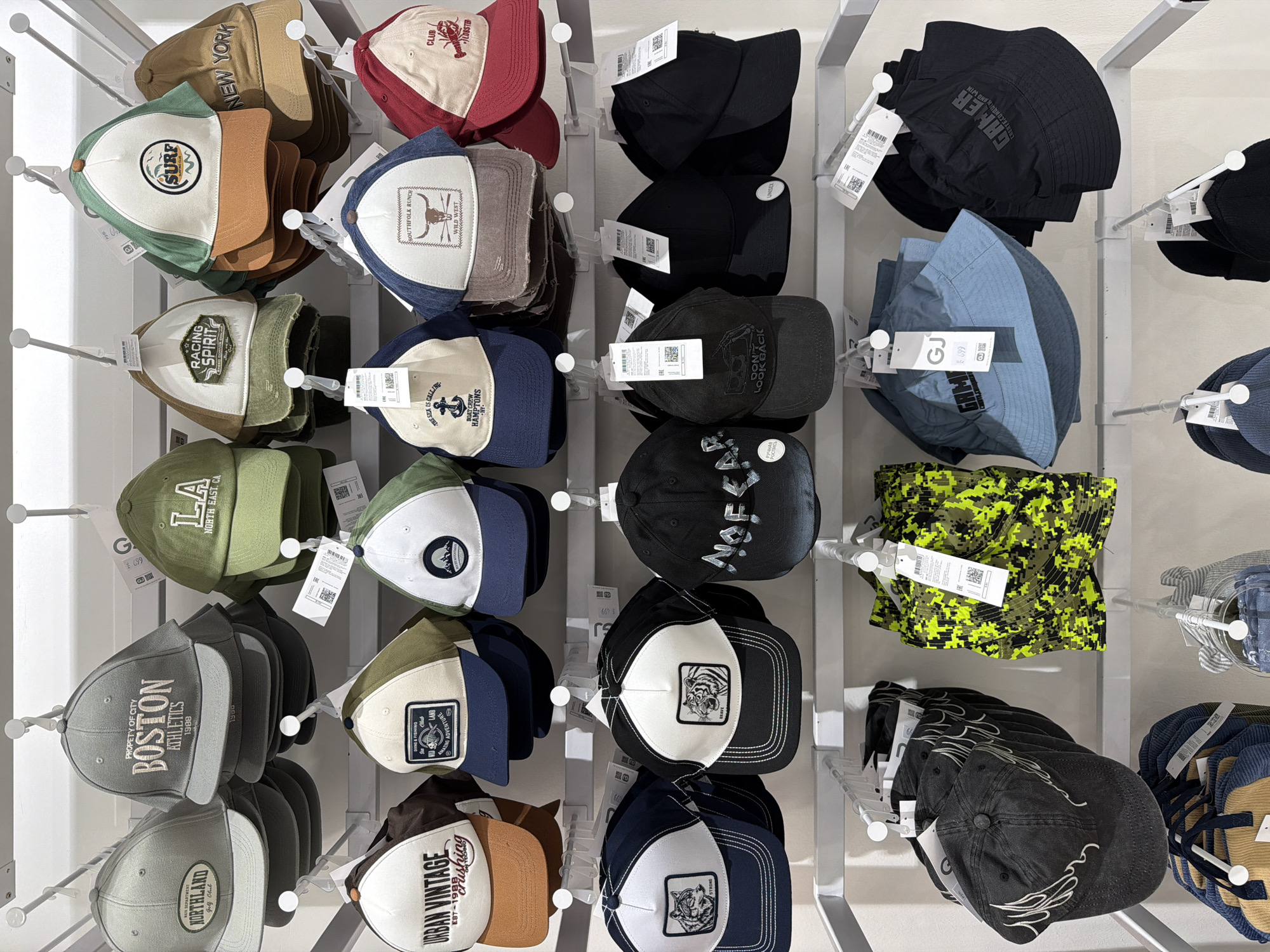 The image displays a well-lit retail environment, likely a section of a store in Chaikovsky, Russia, dedicated to headwear. Multiple shelves or racks are filled with a diverse collection of caps and bucket hats, stacked and displayed for sale. No people are visible; the focus is entirely on the merchandise.
The hats come in various styles, including baseball caps, trucker caps with mesh backs, and bucket hats. A wide range of colors is present, such as khaki, olive green, black, red, blue, grey, white, and brown. Many caps feature embroidered or patched logos and text, indicating different themes or brands.
Notable details include:
* **Logos/Text on Hats:** "NEW YORK," "SURF," "RACING SPIRIT," "LA NORTH EAST. CA," "PROPERTY OF CITY BOSTON ATHLETICS 1988," "NORTHLAND Golf Club," "URBAN VINTAGE EST-19_ _ _ crushing," "THE SEA IS CALLING BOAT CREW HAMPTONS NY," "SOUTHFOLK RANCH WILD WEST," "CLUB LOBSTER," "DON'T LOOK BACK" with a camera icon, "GAMER CREATE. CONQUER AND WIN," and "WILD." Some black caps feature white tiger head patches.
* **Bucket Hats:** A prominent digital camouflage bucket hat in yellow and green is visible, alongside black, blue, and beige ribbed bucket hats.
* **Display:** The hats are neatly arranged on white metal display fixtures, stacked horizontally or vertically, often with multiple hats of the same design grouped together.
* **Pricing/Information:** Many hats have white tags attached, displaying text such as "GJ 699" (likely a price in Russian Rubles), "UNISEX," QR codes, and other product information, including what appears to be "EAC" (Eurasian Conformity) marks.
The scene suggests a modern retail setting, with a focus on casual and sporty headwear.
