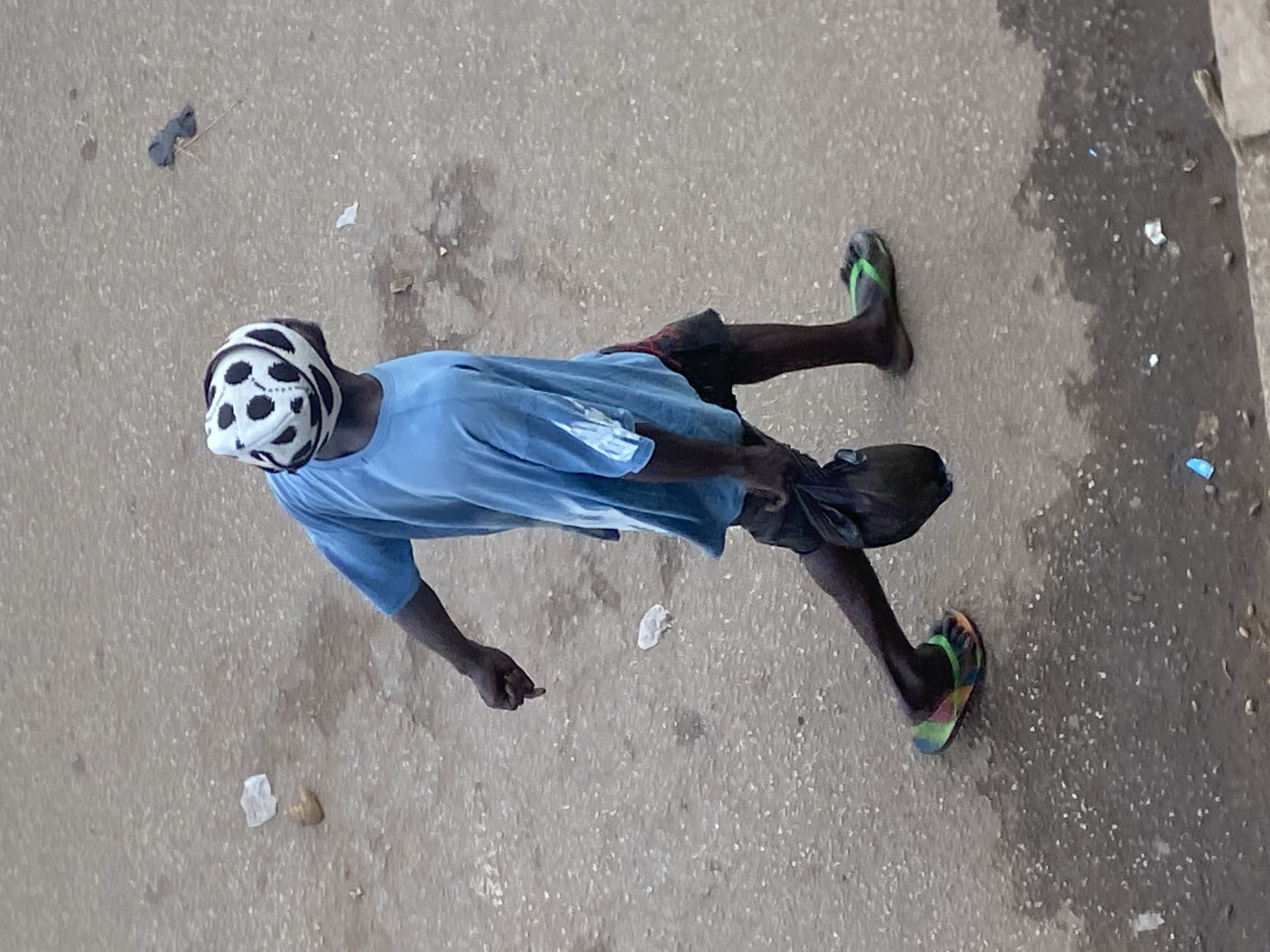 An individual wearing a blue t-shirt, dark shorts, and a white and black patterned headwrap walks on a wet, gritty asphalt surface. The individual is carrying a dark plastic bag in their left hand. Their right hand is extended with the index finger pointing downward. The scene appears to be an outdoor public space in Bukuru, Nigeria, with scattered debris on the ground, including pieces of white paper and a grey cloth. The lighting suggests it is daytime.