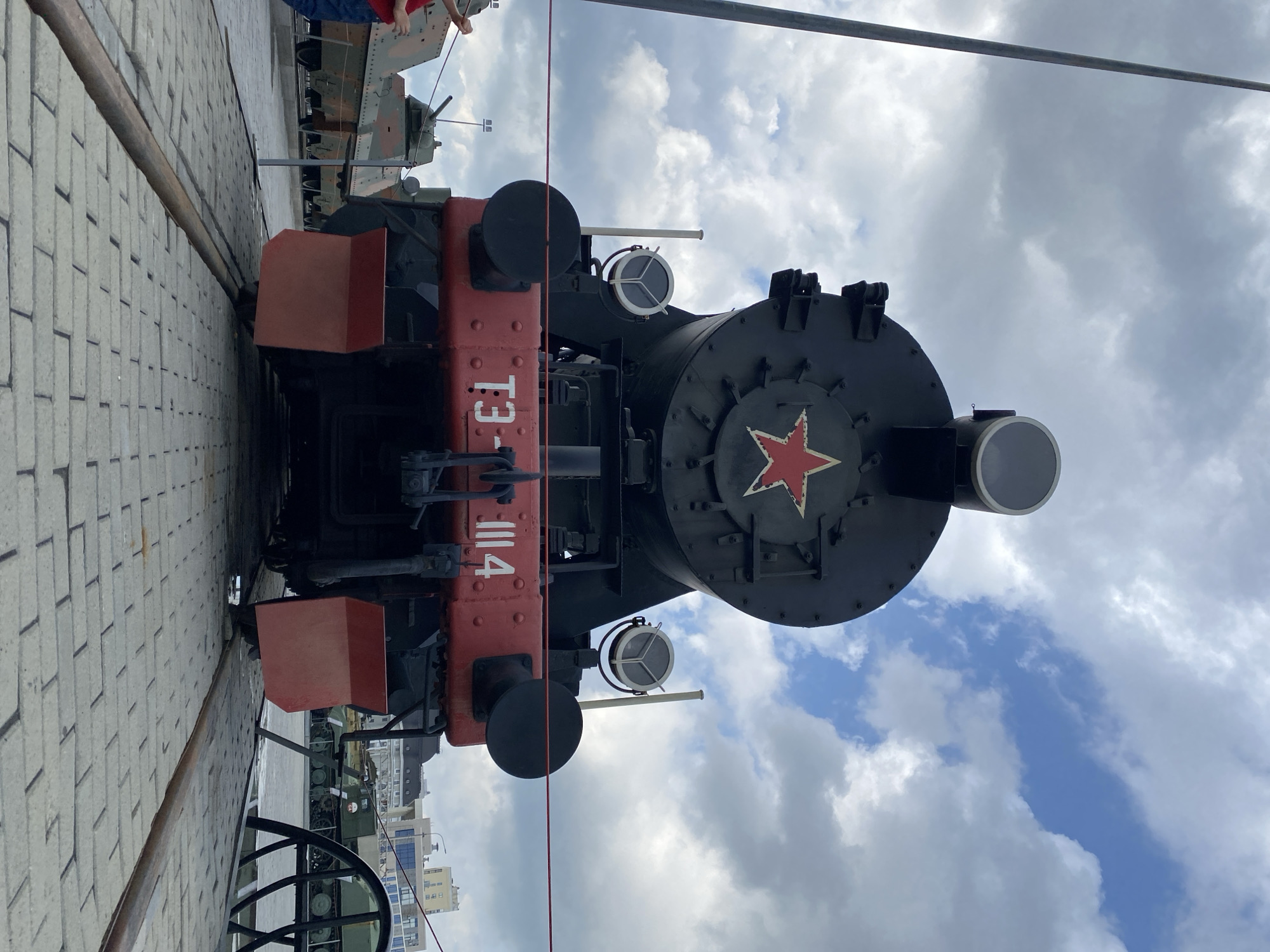 This is a frontal, eye-level, medium shot of a black steam locomotive with a red front bumper, located in the Museum of Military Equipment in Verkhnyaya Pyshma, Russia. The locomotive has a large red star with a white outline on its front. Two circular headlights are mounted above the bumper. The railway tracks in front of the locomotive are made of large grey paving stones. Behind the locomotive, several tanks and military vehicles are parked, some partially obscured by the locomotive itself. In the background, there are buildings and a cloudy sky with patches of blue. A red rope barrier is visible in front of the locomotive, and to the far left, a person in a red shirt and blue jeans is partially visible, looking at the tanks. The overall environment appears to be an outdoor museum or exhibition space.