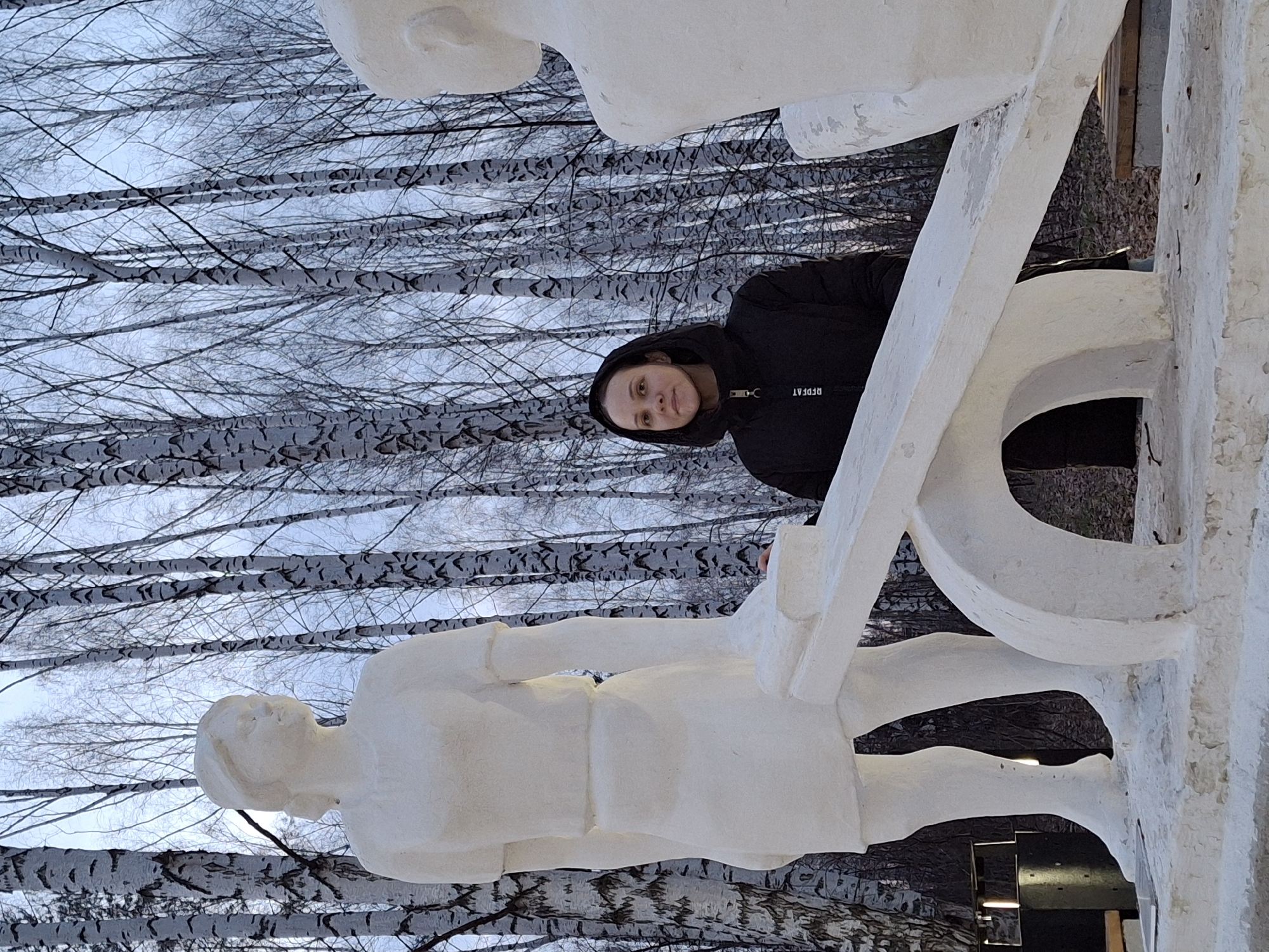 A vertical shot captures a hooded figure standing behind a white concrete sculpture, set against a backdrop of bare birch trees. The sculpture features two human figures, one upright at the bottom and another above, interconnected by horizontal and curved white elements that suggest a ladder or framework. The hooded person is centrally positioned, with the white structure surrounding them, their gaze directed forward. The environment consists of a dense grove of birch trees with white bark and dark markings, their branches intertwining against a light sky. The lower right reveals a glimpse of a modern building facade with dark surfaces and a reflective element. The image conveys a cold, possibly winter, setting. The sculpture is located in the city of Chaikovsky, Russia.