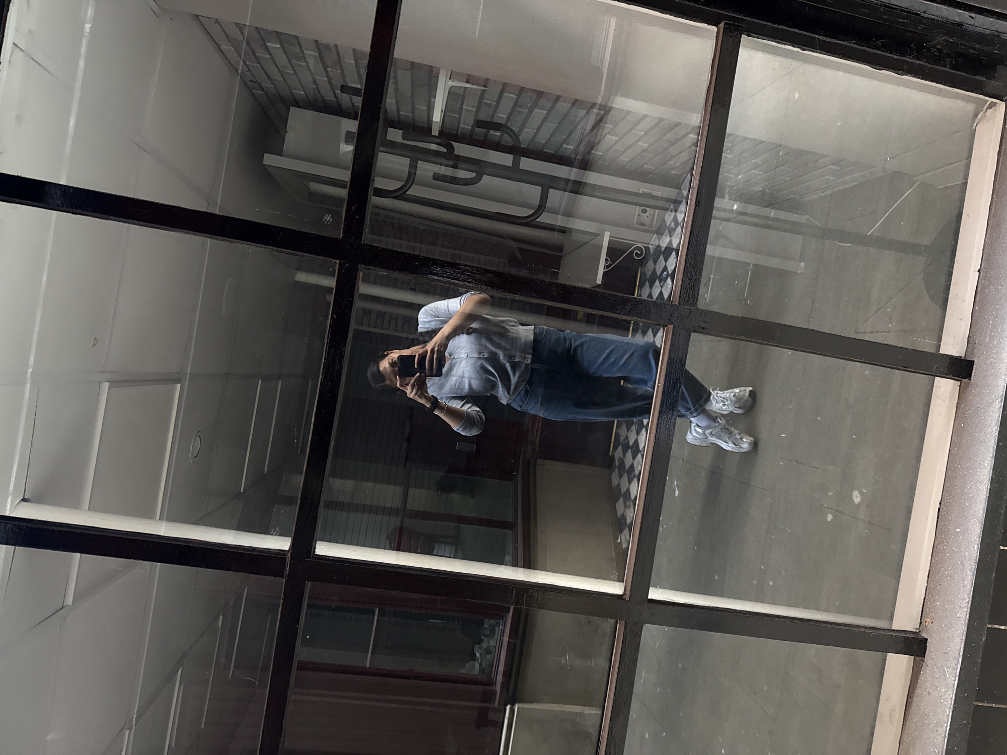 The image depicts a reflection of a person taking a photograph of themself in a multi-pane window. The window frame is black. The person is wearing a light-colored top and blue jeans and has on silver-colored shoes. The reflection reveals the setting to be a building with white walls, black and white checkered tiles on the floor, and visible pipes or fixtures on the ceiling. The environment appears to be indoors. There is no way to determine the location within Great Yarmouth from this image alone.