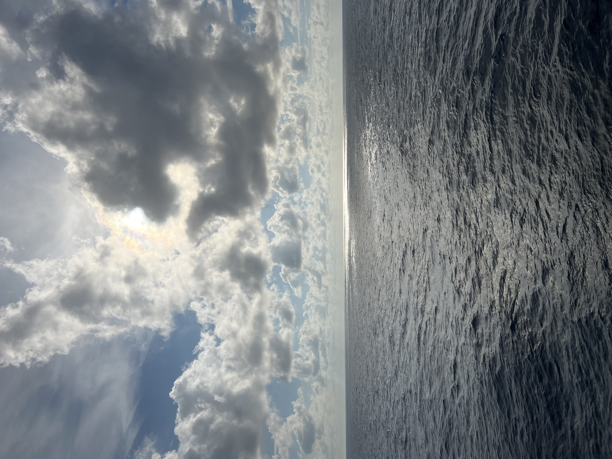 The image captures a vast expanse of the ocean under a dramatic sky. The water is a deep, dark blue, with ripples and waves reflecting the light. A bright, shimmering band of sunlight stretches across the water's surface, creating a path of reflected light. Above, the sky is a mix of blues and whites, with large, puffy clouds casting shadows. Some of the clouds are illuminated by the sun, creating a soft, ethereal glow. A subtle rainbow or prismatic effect is visible near the sun, peeking through a gap in the clouds. The scene evokes a sense of immense scale and natural beauty. The location is stated as Unknown, Unknown, with no specific city details provided.