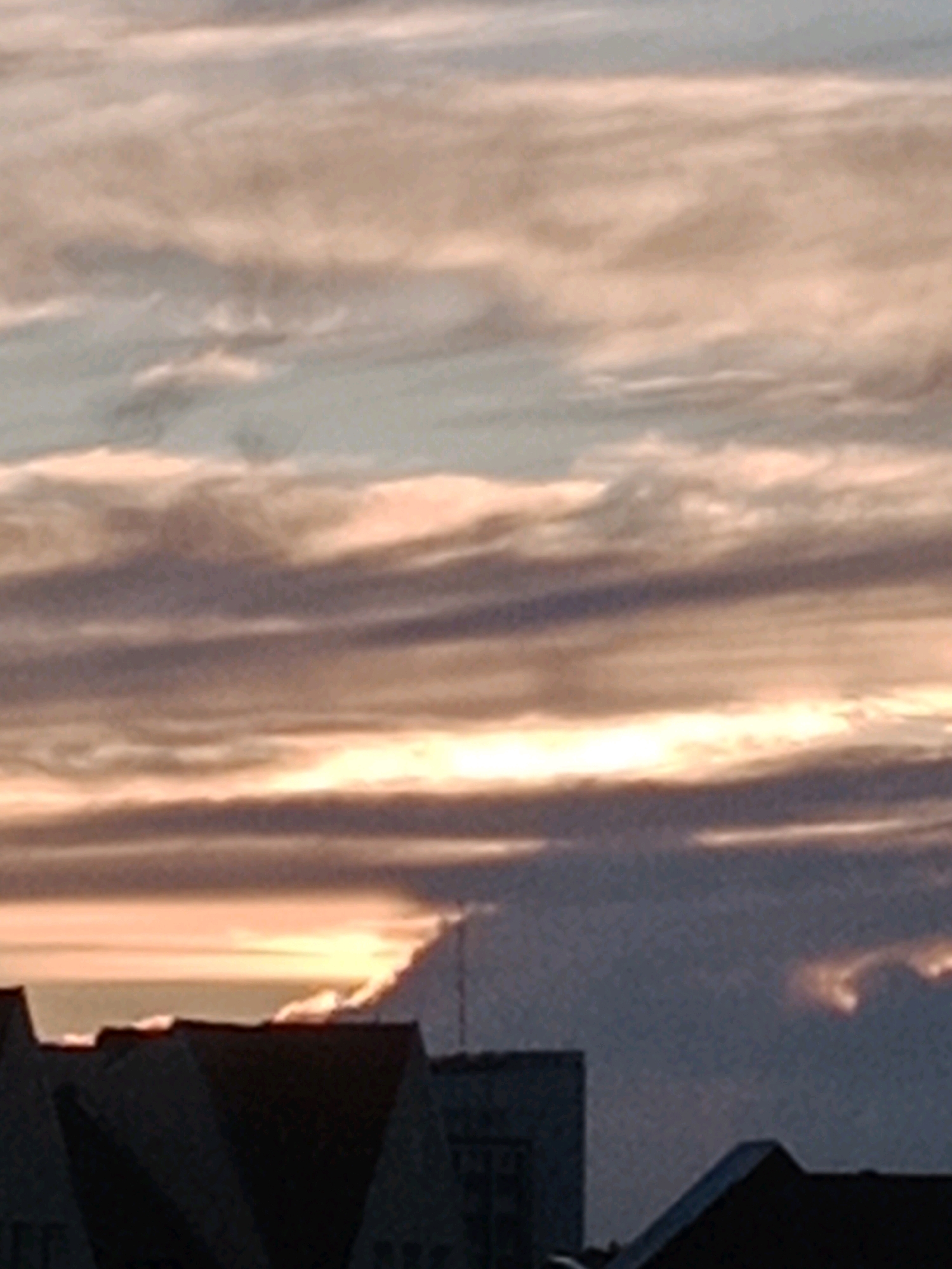 The image shows a silhouetted cityscape beneath a dramatic sky. The foreground is dominated by the dark shapes of building rooftops, with at least two distinct rooflines visible. In the background, the sky is filled with layered clouds, exhibiting a gradient of colors from pale orange and yellow near the horizon to darker blues and grays higher up. The lighting suggests either a sunrise or sunset. The overall scene is devoid of any people or overt activity, presenting a static urban landscape against a dynamic sky. The location is identified as Bydgoszcz, Poland.