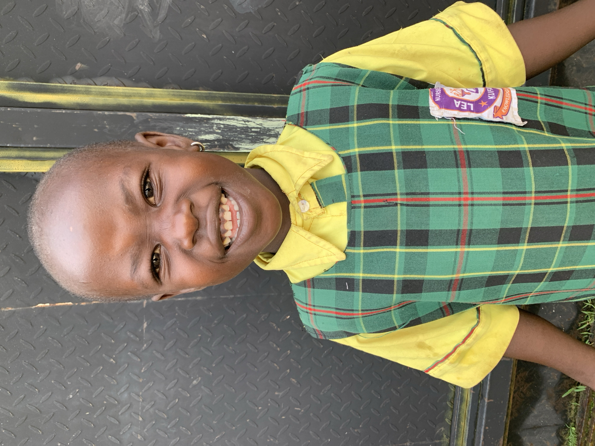 A young child, with dark skin and a shaved head, smiles broadly at the camera. The child is wearing a yellow collared shirt with a green and black plaid vest over it, which has a white and purple patch on the left side. The patch contains an emblem with the words "LEA KUFARA" and "NURSERY KNOWLEDGE." The child has a silver hoop earring in their left ear. The background is a dark, textured metal surface with vertical yellow lines. The scene appears to be outdoors, with some green vegetation visible at the bottom. The child's facial expression is joyful, with visible teeth and crinkled eyes. The lighting suggests it is daytime, with a slight shadow on the child's face. The overall impression is one of a happy child in a school uniform.