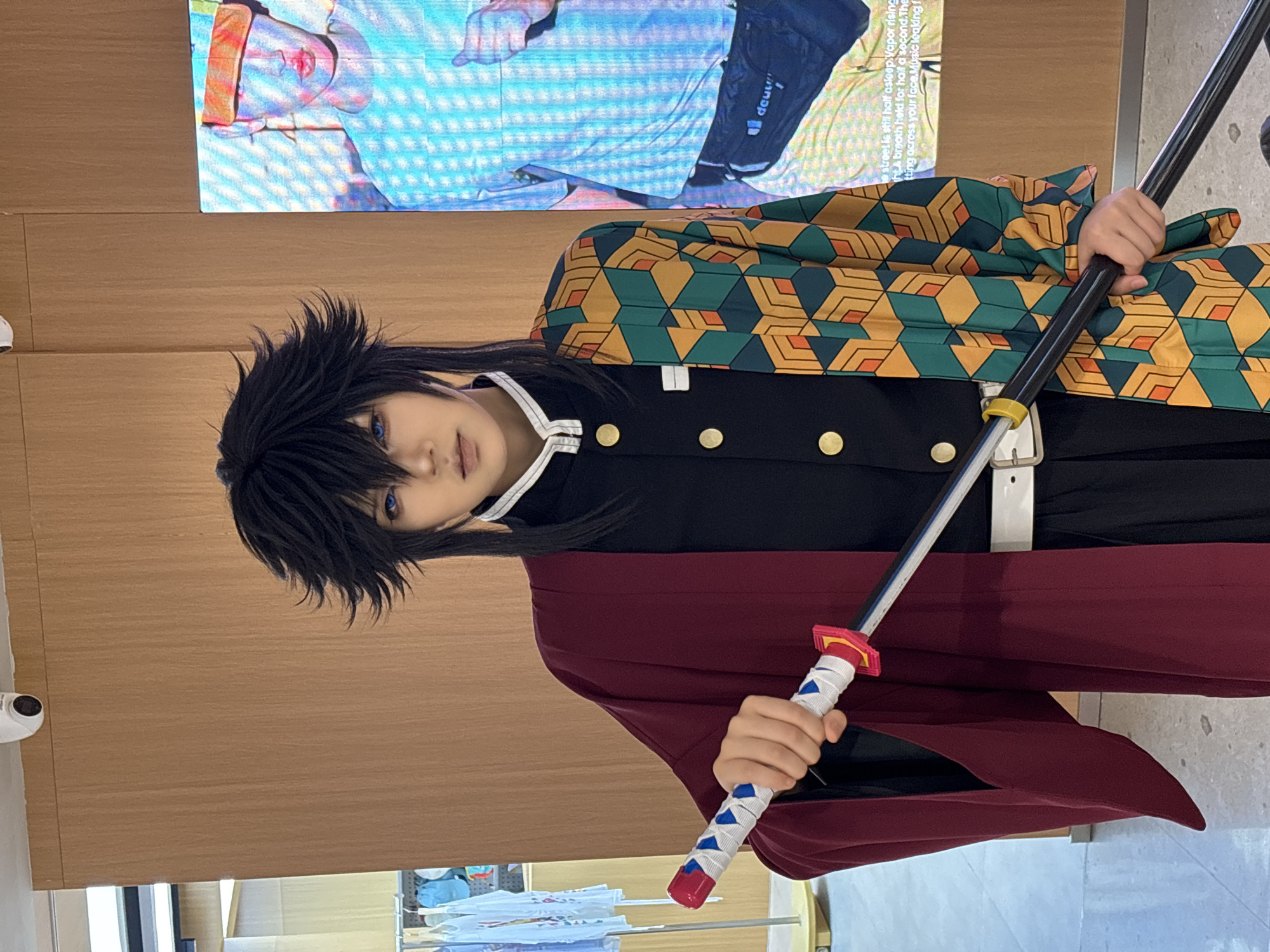 A person dressed as Giyu Tomioka from the anime series Demon Slayer holds a sword with a white handle and a blue and red wrapping. The person wears a black uniform, a green and orange patterned haori, and a maroon cloak. They have black, spiky hair and blue contact lenses. The scene is indoors, in a public space, with wood-paneled walls and a monitor showing a person wearing athletic wear. The photo was taken in Liangjiang, China.