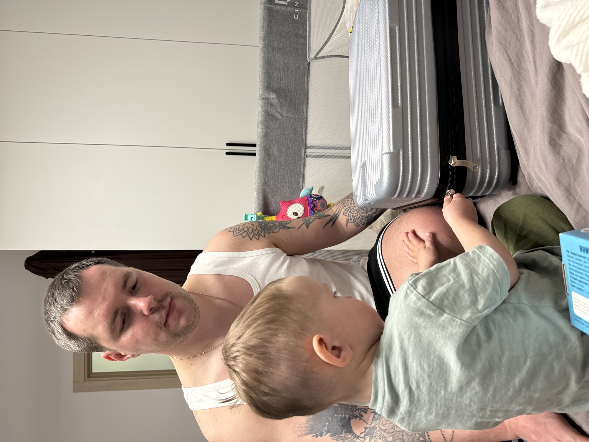 A man and a toddler are together indoors. The man is fair-skinned with short, graying hair and a gold chain necklace, wearing a white tank top. He has prominent tattoos on his arms. A young child with blond hair is sitting close to him, wearing a light green t-shirt. The child's hand is touching a silver suitcase. Next to the suitcase there is also a baby cot with a gray frame. A colorful toy is resting near the man's leg. The environment includes a white closet, suggesting a bedroom setting. The location is Odintsovo, Russia.