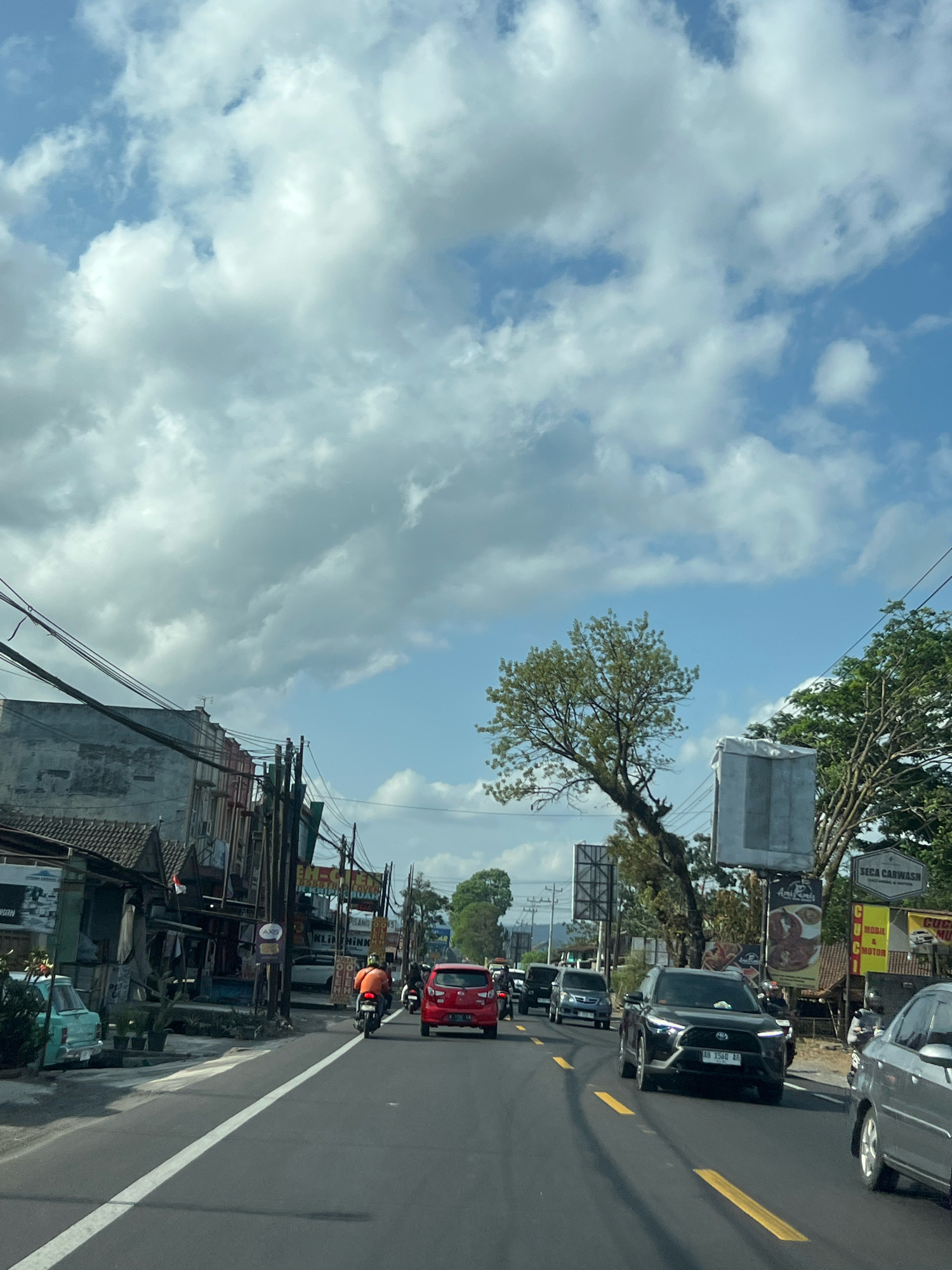 The image depicts a street scene in Jambewangi, Indonesia, with a view of the sky filled with scattered clouds and blue sky. The road is paved with asphalt and marked with white and yellow lines. Several vehicles are present, including a red car, a black SUV, a silver car, and multiple motorcycles. One motorcycle rider, wearing an orange shirt, is visible. Along the side of the road, there are buildings with various signs, including one for "SECA CARWASH". Trees and utility poles with numerous wires are also visible. The overall environment suggests a moderately busy urban or semi-urban area during daylight hours.
