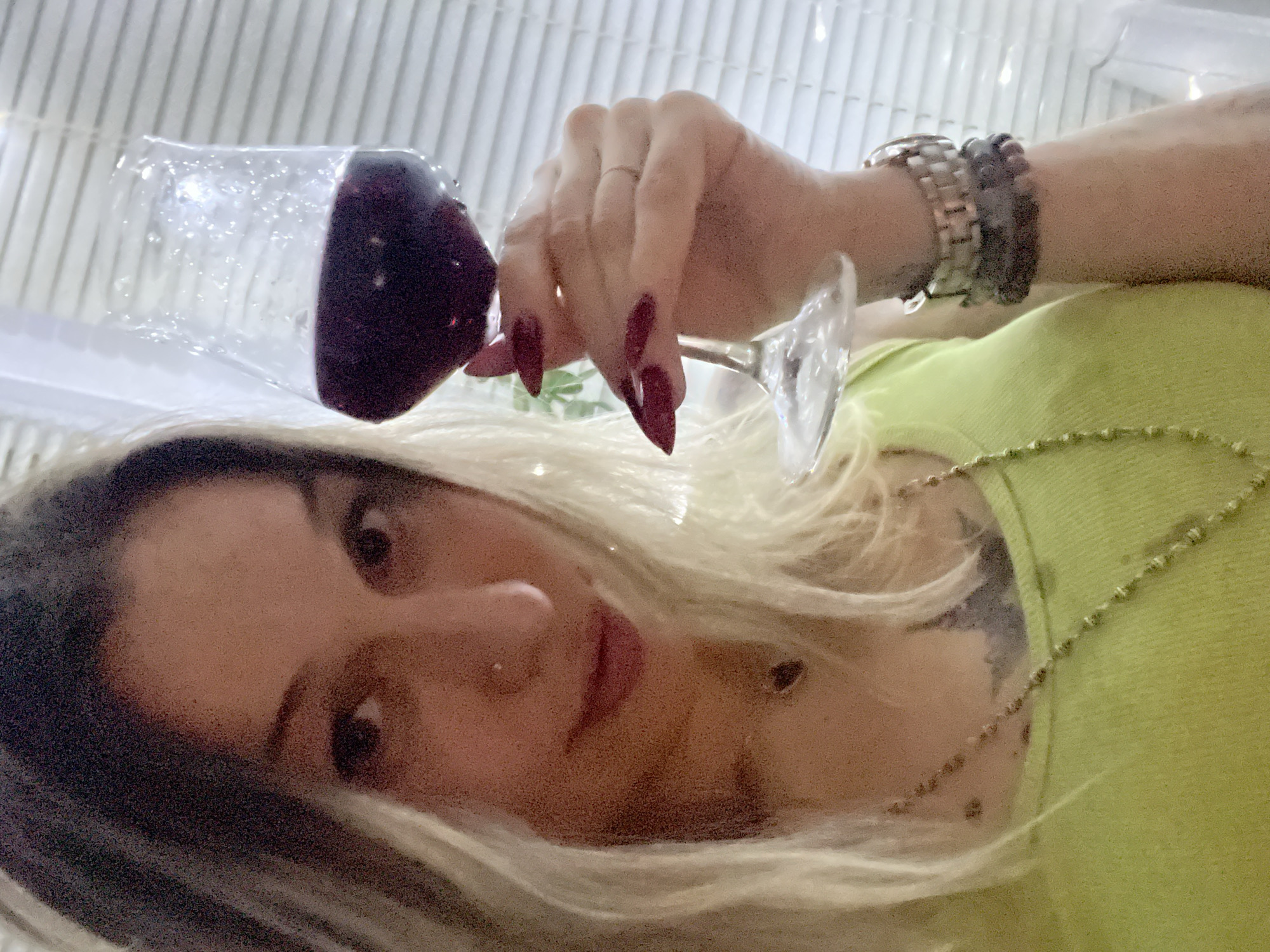 An unknown woman is pictured indoors holding a glass of red wine. She has blonde hair and is wearing a lime green tank top and several necklaces and bracelets. Her nails are long and painted dark red. The background shows a window with blinds and some string lights.