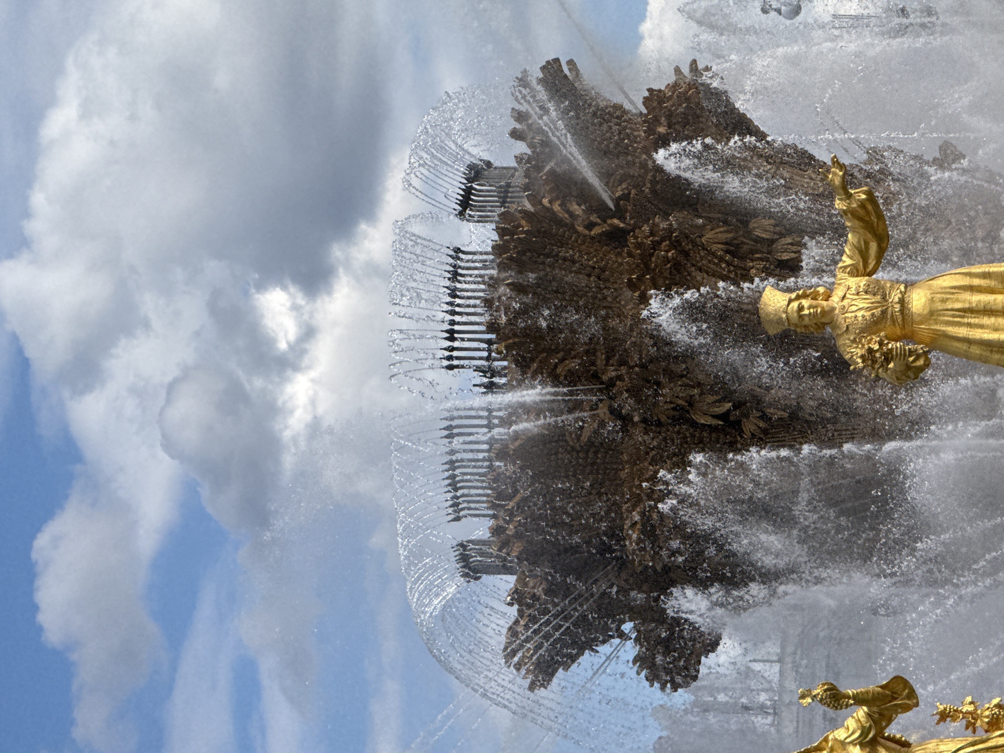 This is a close-up, vertically oriented shot of a large, ornate fountain in Moscow, Russia. The fountain features elaborate golden sculptures and a complex water display. In the foreground, a golden statue of a woman in historical attire, wearing a crown or headdress, is partially visible. She holds a bouquet of what appears to be flowers or fruit in her left hand and her right arm is raised. Behind and above her, the main structure of the fountain is a mass of textured, rock-like formations from which water cascades and sprays. Numerous thin streams of water arc outwards from the central structure, creating a shimmering curtain effect against the sky. A second, smaller golden sculpture, possibly a cherub or another figure holding grapes, is visible in the lower right corner.
The sky in the background is a mix of bright blue and dramatic, heavy clouds, suggesting potentially changeable weather, perhaps a partly cloudy day. The sunlight catches the water, creating a bright, dynamic scene. The overall impression is one of grandeur and movement, highlighting the artistry of the fountain and the power of the water. The detailed craftsmanship of the golden sculptures and the intricate design of the water jets are prominent. The scene appears to be set outdoors, likely in a park or public square, given the scale of the fountain and its prominent placement. No visible text is present in the image.