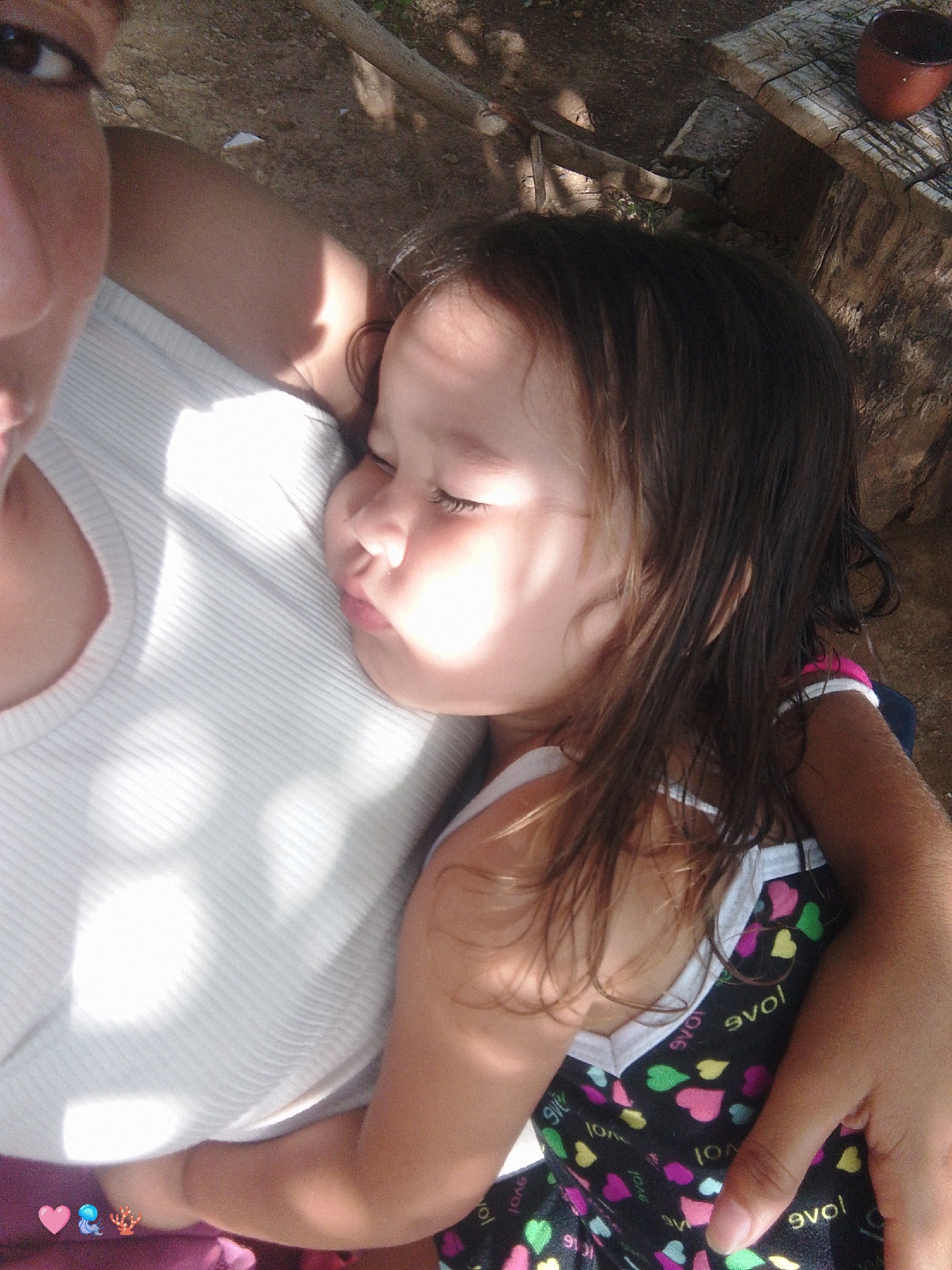 The image is a close-up, overhead shot of an adult holding a young child. The adult's left arm is visible, supporting the child who is nestled against the adult's chest. The child appears to be asleep, with eyes closed and head resting on the adult's shoulder. Both individuals are wearing casual clothing. The child has dark hair and is wearing a patterned garment with hearts. The adult is wearing a white, ribbed top. The background is out of focus but suggests an outdoor setting with natural elements such as dirt, wood, and possibly foliage. The lighting indicates a sunny day, with shadows falling across the scene. The image may have been taken in Santa Ana, Venezuela, based on the context provided.