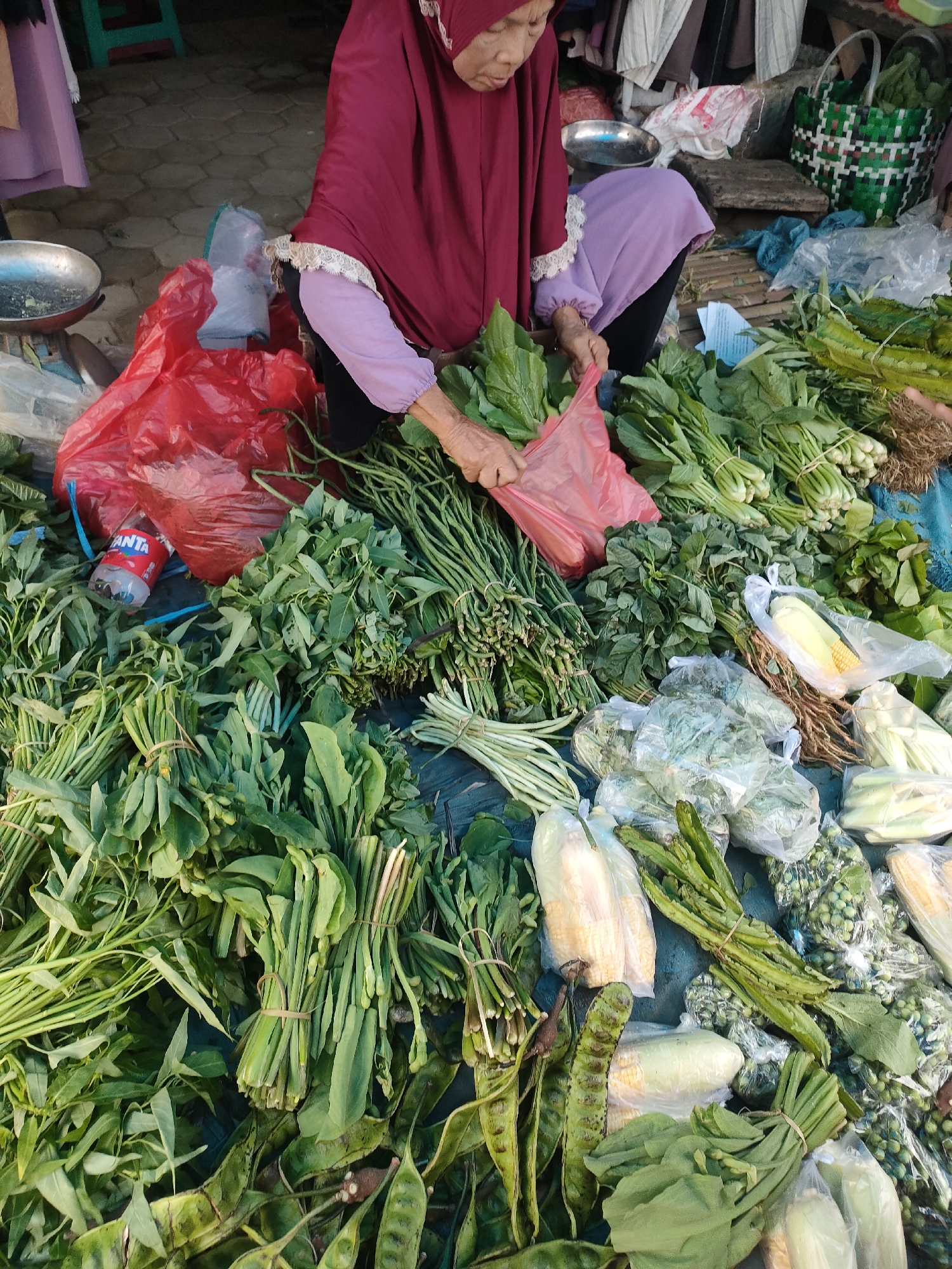 This is a close-up, eye-level, outdoor, medium shot of a woman selling vegetables in the Sukoyoso market, Indonesia. The woman is wearing a maroon hijab and a purple garment, and she is seated on the ground. She is in the process of bagging vegetables. She is surrounded by a large assortment of fresh vegetables, including bundles of leafy greens, long beans, corn, and other produce. The vegetables are arranged on the ground and on a dark blue tarp. There are also red plastic bags, a metal scale, and a green woven basket visible in the background. The lighting suggests it is daytime.