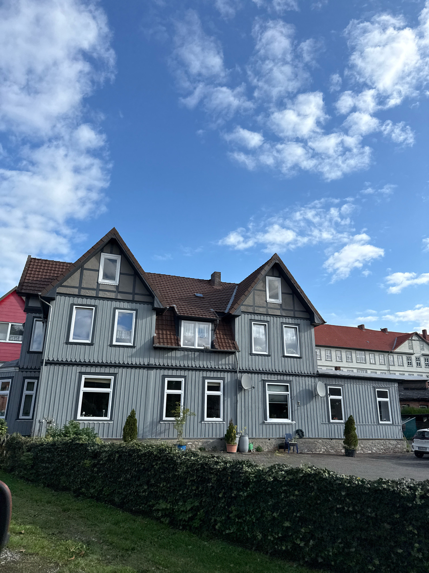The image shows a multi-story building with a gray, vertical wooden facade, common in historical German architecture. The building features numerous windows with white frames. The roof is tiled in a reddish-brown hue. In the foreground, a dense green hedge runs along the front of the property, with a grassy area beyond. The sky above is a vibrant blue, dotted with scattered white clouds. To the right, a white car is parked in a driveway. The architectural style and the presence of what appears to be a historical building suggest a location within a European town or city. Based on the provided context, this scene is located in Osterode am Harz, Germany, a town known for its well-preserved half-timbered houses and historic buildings. The image captures a residential or commercial property on a clear day.