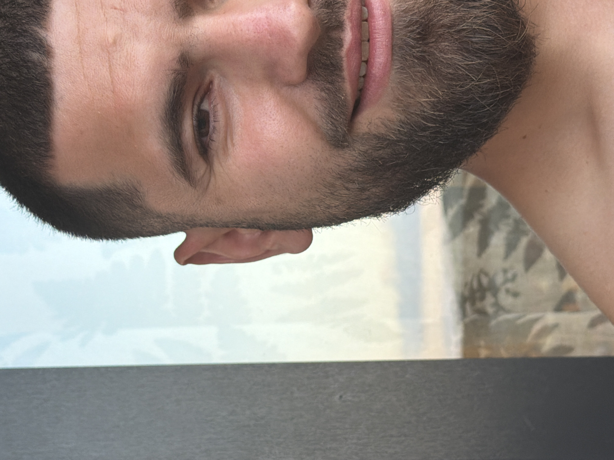 A close-up, cropped, portrait-style image shows the right side of a man's face. He has a short buzz cut and a full beard, with a closed-mouth smile revealing his teeth. His right ear is visible. The background consists of a blurred, light-colored pane of glass with a hint of sky and some faint patterns behind it. The left side of the image is dominated by a dark, textured surface. The lighting suggests it is daytime. The image does not provide any specific information about the location in Florești, Romania, or any actions or interactions.