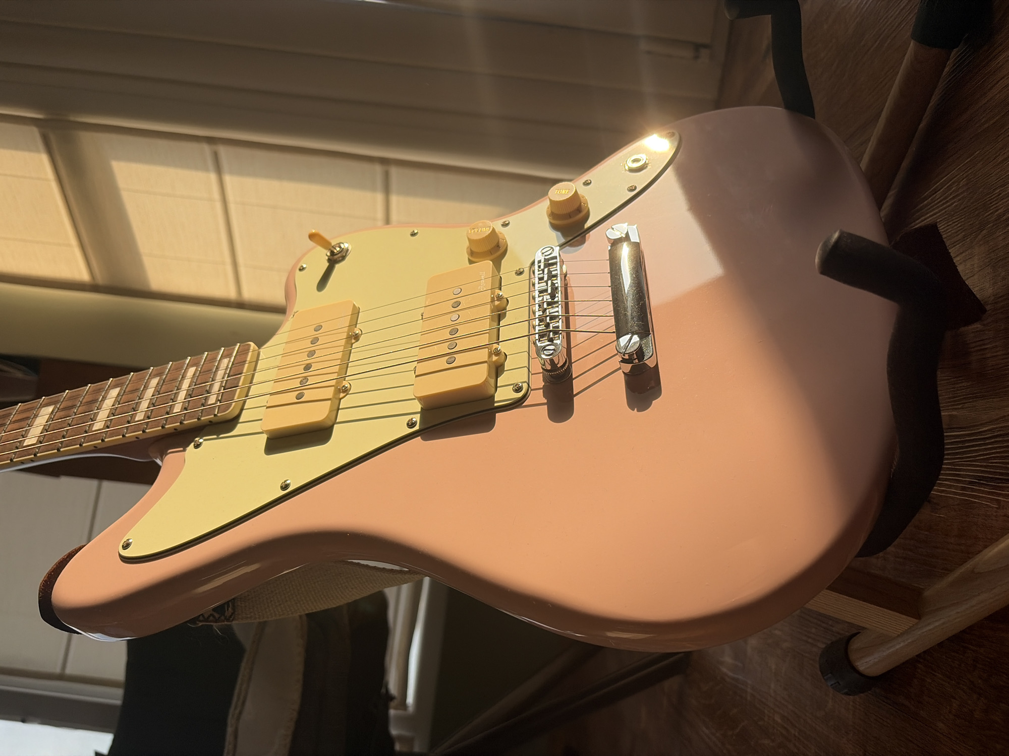 An electric guitar, featuring a pink body and a cream-colored pickguard, stands on a wooden guitar stand. Two cream P90-style pickups are mounted on the pickguard, along with two cream control knobs labeled "VOLUME" and "TONE" and a three-way toggle switch with a cream tip. A chrome bridge and vibrato system are present on the body. A beige fabric strap is attached to the guitar's upper horn. The guitar neck, with a visible fretboard and rectangular inlays, extends upwards. The guitar stand consists of a light-colored wooden frame with black rubber padding. The floor beneath is composed of wooden floorboards with a visible grain. Strong natural sunlight enters from the right side of the frame, illuminating the guitar and casting distinct shadows across its body and the floor. In the background to the right, a window with horizontal blinds or slats is visible, partially covered by light-colored curtains. To the upper left, a portion of dark-colored furniture is visible. No individuals are present in the scene.