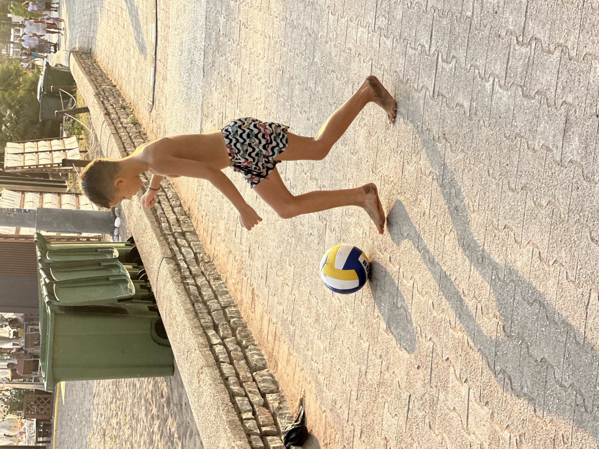 A young boy, shirtless and wearing patterned swim trunks, plays with a volleyball on a paved walkway in Boreti, Montenegro. He is positioned in the center of the frame, facing right, with his left leg extended and his right leg bent as if kicking the ball. The volleyball, a blue, yellow, and white patterned sphere, sits on the ground in front of him. His feet are dirty, suggesting he has been playing on the nearby sandy beach.
The walkway is made of interlocking paver stones with a textured surface, and long shadows are cast across it from the setting sun. To the left of the walkway, a low, rough-hewn stone wall separates it from a sandy area. Behind the wall, several large green trash bins are lined up. In the background, a blurred scene of beachgoers, tables, chairs, and vegetation suggests a public area near the beach. Further in the distance, more people are walking along a path. The overall scene is illuminated by the warm, golden light of late afternoon or early evening.
