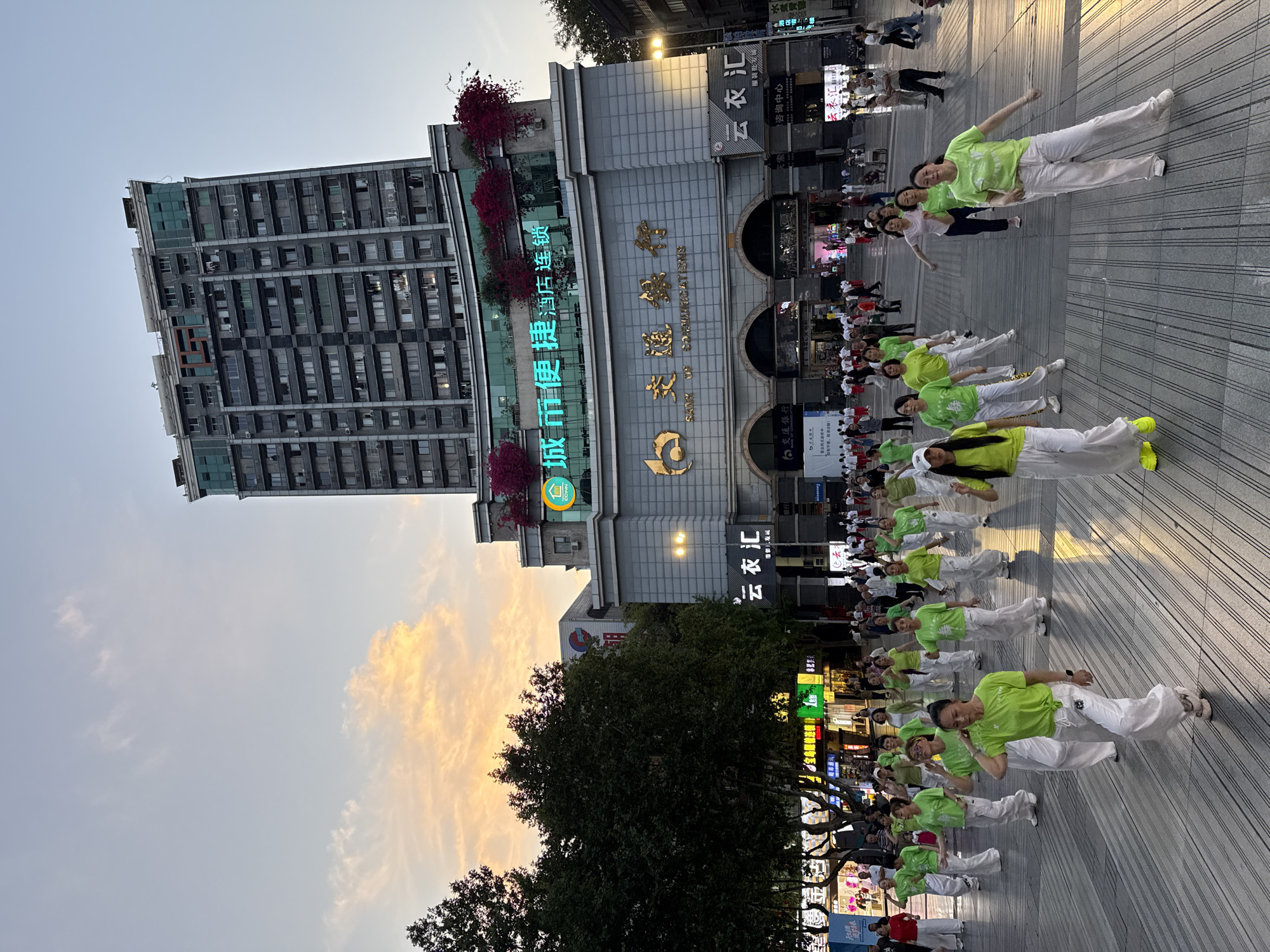 The image depicts a group of women dressed in matching green shirts and white pants performing synchronized exercises in a public square in Beibei, China. The dance group is spread across a pedestrian area with a grooved surface. In the background is a building with Chinese characters and flowers. Other people are seen observing the exercise. The sky in the background has a sunset glow. Commercial signs, including those for "云衣汇" and "交通銀行," are visible. A tall building towers in the background to the left.