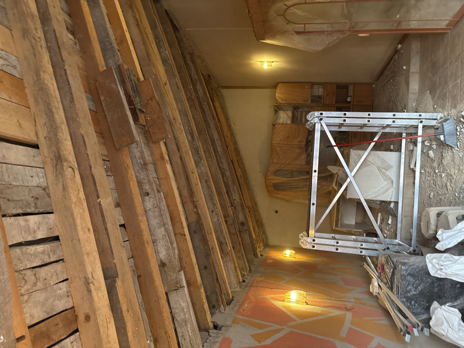 The scene depicts an interior room or space, possibly under renovation or used for storage, located in Stefan Karadja, Bulgaria. The sloped ceiling is constructed from numerous exposed wooden planks of varying dimensions and finishes, arranged in a rustic manner with visible gaps.
The left wall features a geometric design in shades of orange and a lighter color. Two wall-mounted sconces with textured glass covers illuminate this wall with a warm yellow glow. The rear and right walls are painted a uniform, light, neutral color, with one identical wall sconce illuminating the right wall.
In the central foreground, an erected silver-colored aluminum scaffolding structure is prominent. A red-handled broom with black bristles leans against the right side of the scaffolding. The floor on the left side and beneath the scaffolding is composed of dirt, dust, and construction debris. This ground transitions to a lighter-colored tiled floor on the right side, separated by a slight elevation or step.
Along the back wall, multiple items of wooden furniture, including cabinets and shelving units, are positioned and uniformly draped with translucent plastic sheeting, indicating protection from dust or temporary storage. A larger, indistinct object, also covered in plastic, is visible further left along the back wall. In the lower-left corner, various wooden planks, fragments of building material, and a black tarp-covered item are arranged. Two small, white, plastic stools are partially visible in the bottom-left foreground. No individuals are present in the image.
