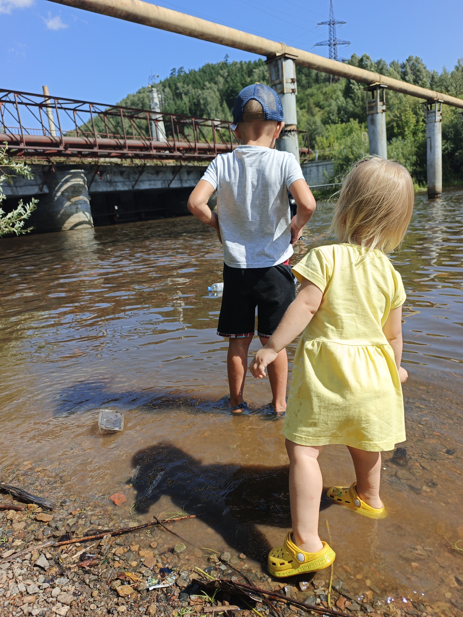 The image depicts two children, a boy and a girl, standing in shallow, muddy water near a rocky shoreline. A bridge, possibly an aqueduct, crosses the water in the background, supported by concrete pillars. Trees and a power line tower are visible on the far bank. The boy, wearing a blue cap and grey t-shirt, stands facing away from the camera, looking towards the bridge. The girl, in a yellow dress, is also facing away, her attention seemingly directed towards the water. A clear plastic container floats near the shore, and various pieces of debris, including sticks and litter, are scattered on the bank. The water reflects the sunlight, creating ripples and glints. The scene appears to be an outdoor recreational area near a body of water, possibly a river or reservoir, within the city of Zlatoust, Russia, given the prominent industrial infrastructure and natural landscape.