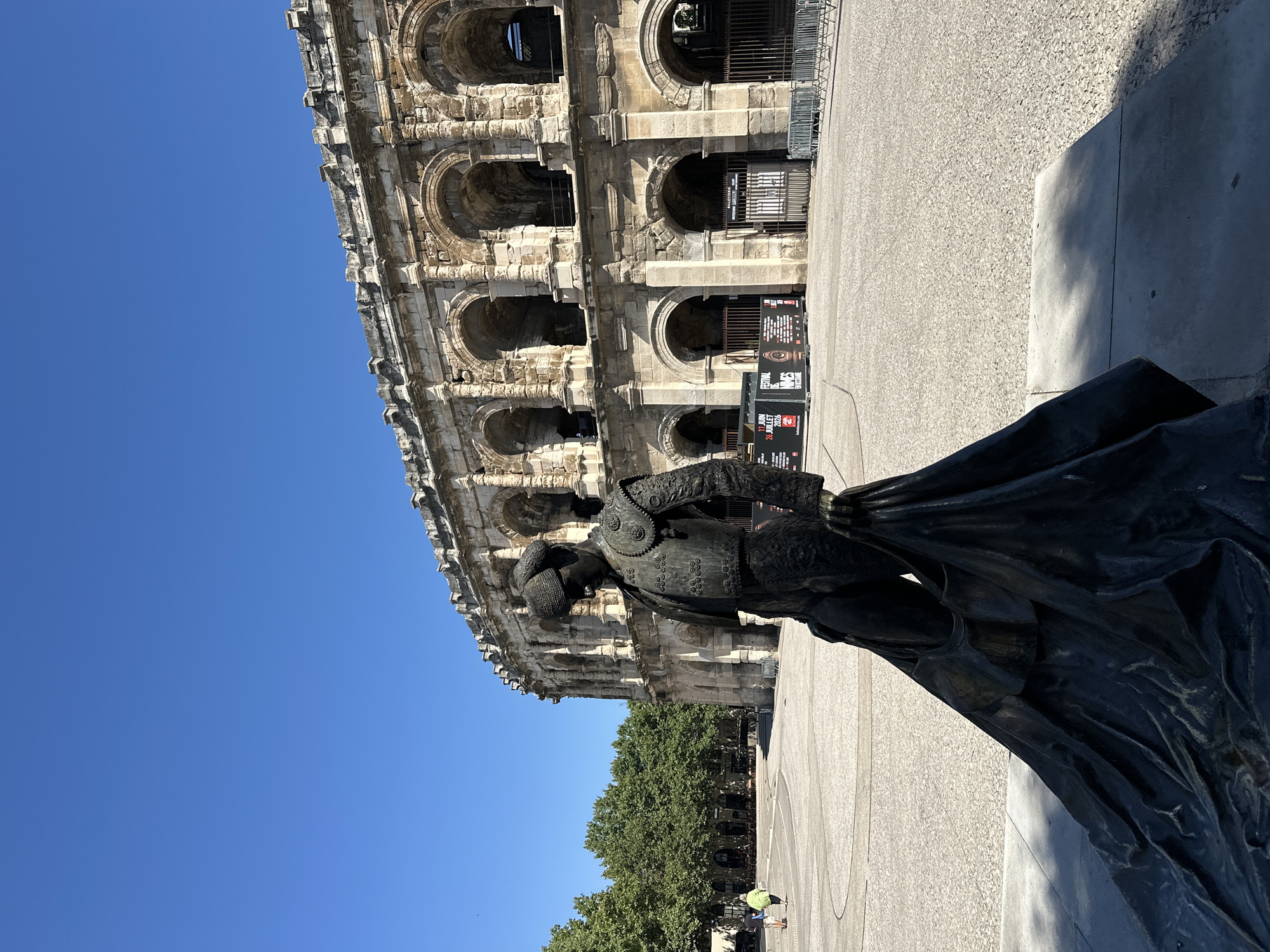 A bronze statue of a bullfighter stands in front of a large, ancient stone structure with arched openings. The bullfighter is depicted in traditional attire, including a cap, ornate jacket, and a flowing cape. The stone structure appears to be a Roman amphitheater, with multiple levels of arches. In the background, lush green trees line a pathway, and several small figures of people can be seen walking. The sky is a clear, bright blue, suggesting a sunny day. The overall scene is one of historical significance and cultural heritage. A poster near the amphitheater entrance displays the text "FESTIVAL NÎMES 2023" and "11 JUIN 26 JUILLET". The statue is positioned in a way that it partially obscures the view of the amphitheater, creating a sense of depth and perspective. The lighting indicates it is daytime, likely midday given the strong shadows.