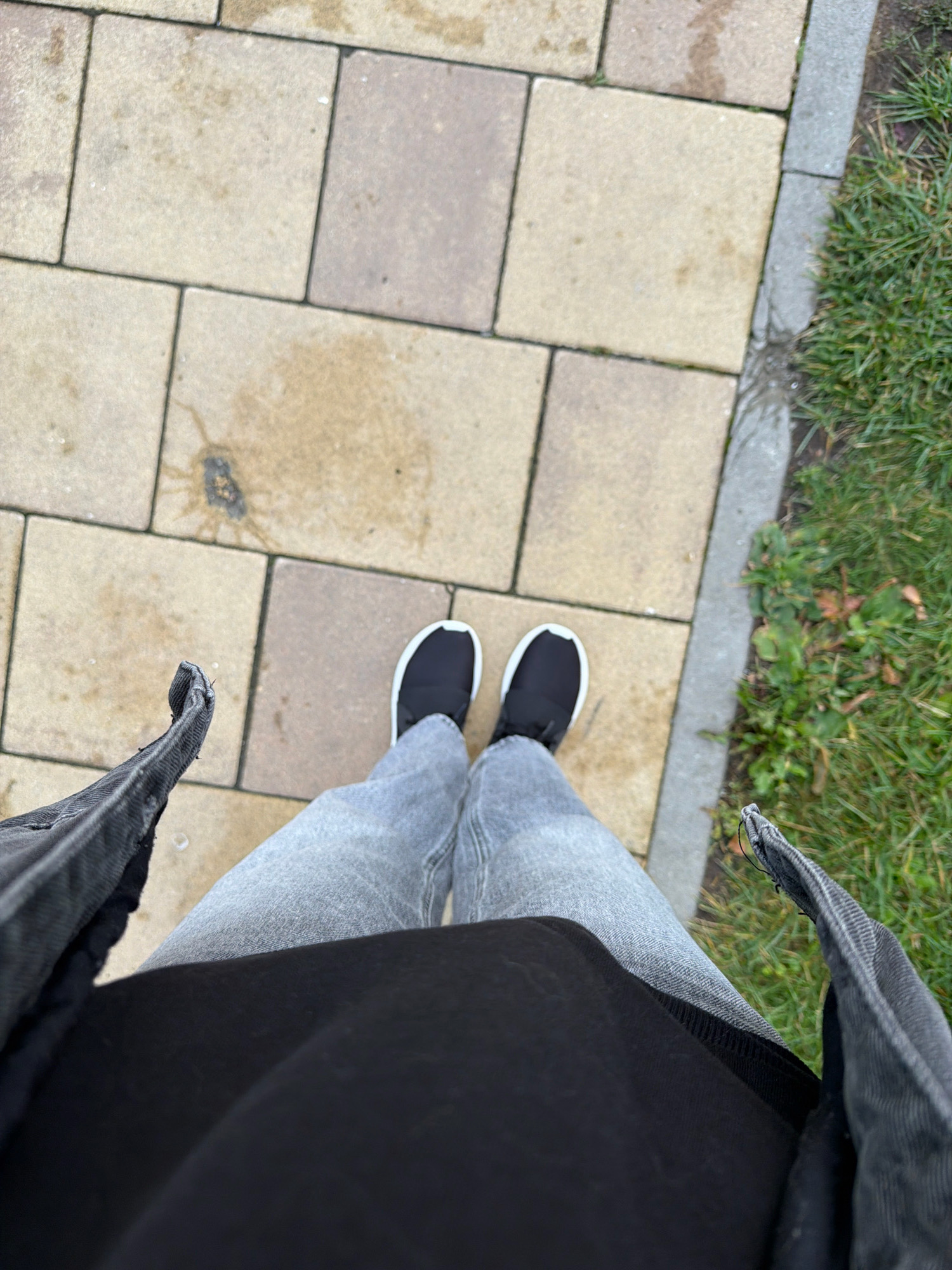 An overhead shot shows a person standing on a paved walkway. The walkway is made of light brown square tiles. To the right of the walkway is a strip of green grass. The person is wearing light gray jeans, black shoes with white soles, a black t-shirt, and a dark gray denim jacket. The person's legs and feet are visible in the lower half of the frame. The scene appears to be outdoors, possibly in a park or residential area. There are no discernible landmarks that would indicate the specific location within Kyiv, Ukraine.