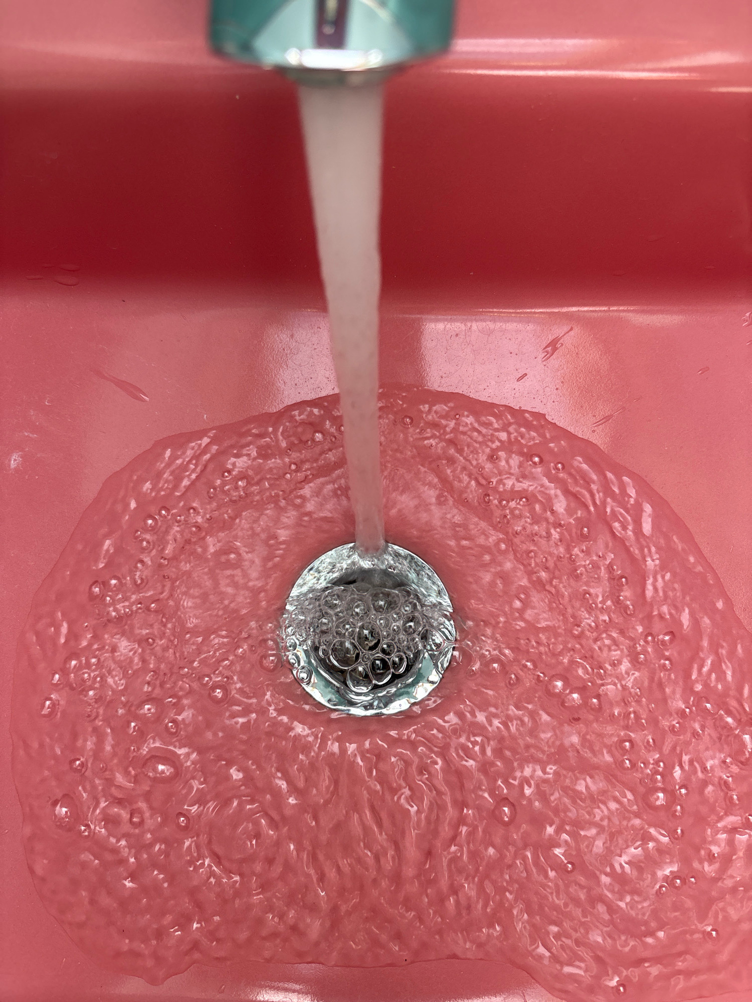 This is a close-up, top-down view of a pink sink with water running from a faucet. The water is a pale white and is flowing directly into a silver drain. The sink basin is filled with a shallow layer of water, which is creating ripples and bubbles around the drain. There are a few small water droplets visible on the surface of the sink. The lighting is bright and even. There are no people or animals visible in the image. The scene is forensic and matter-of-fact, depicting a mundane action in a domestic setting. The image does not contain any information about the location in Reykjavik, Iceland.