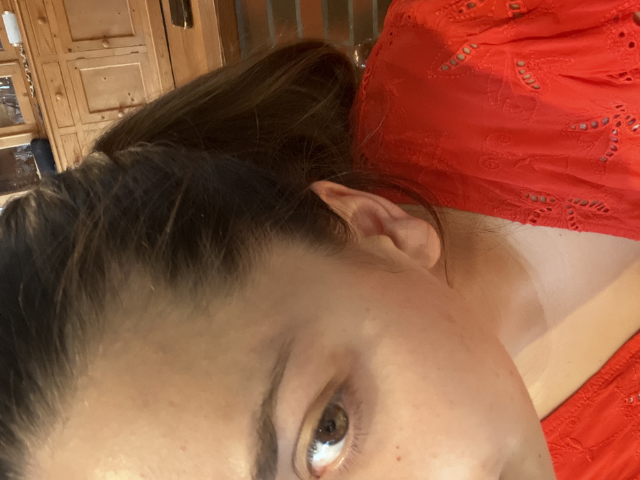 The image displays a close-up, angled view of an individual's right side, identified as being in Ancenis, France. The subject's right eye, with a visible brown iris and dark pupil, is in the lower left quadrant. The forehead, part of the nose bridge, cheek, and right ear are visible. The skin tone is fair with minor imperfections on the forehead and cheek. Dark brown hair, appearing to be pulled back into a ponytail, frames the face and ear; lighter brown strands are discernible within the ponytail. The subject is wearing a bright red garment, likely a top or dress, featuring patterned eyelet cutwork on the visible shoulder and upper arm. The background is out of focus, revealing a light-colored wooden cabinet with multiple drawers and wooden knobs in the upper right. Below the cabinet, a dark horizontal surface, possibly a table or counter, is visible, alongside a reddish-brown textured surface resembling brickwork.