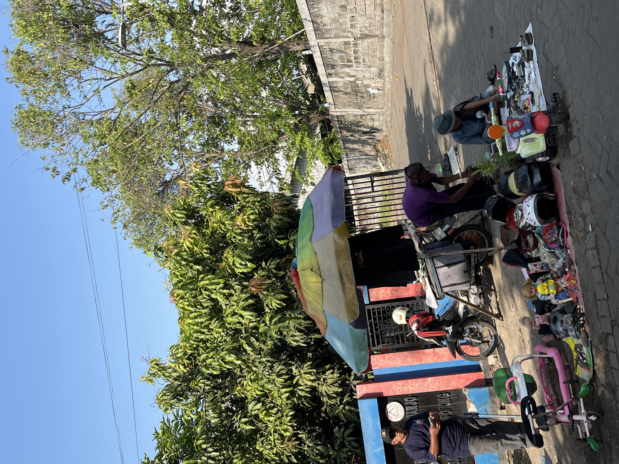 In Kesambi, Indonesia, a street scene unfolds under a clear blue sky, with lush green trees and a stone wall forming the backdrop. In the foreground, a collection of items is laid out on a mat on the paved ground. These include several helmets, a yellow duck toy, dolls, and various small objects. To the left, a man in a dark blue shirt and grey pants stands holding a phone, next to a pink baby walker. A motorcycle is parked behind a colorful umbrella-covered stall, where a person in a purple shirt is attending to the displayed items. Further to the right, another person, wearing a blue shirt and a hat, is seated and sorting through more items on the mat.