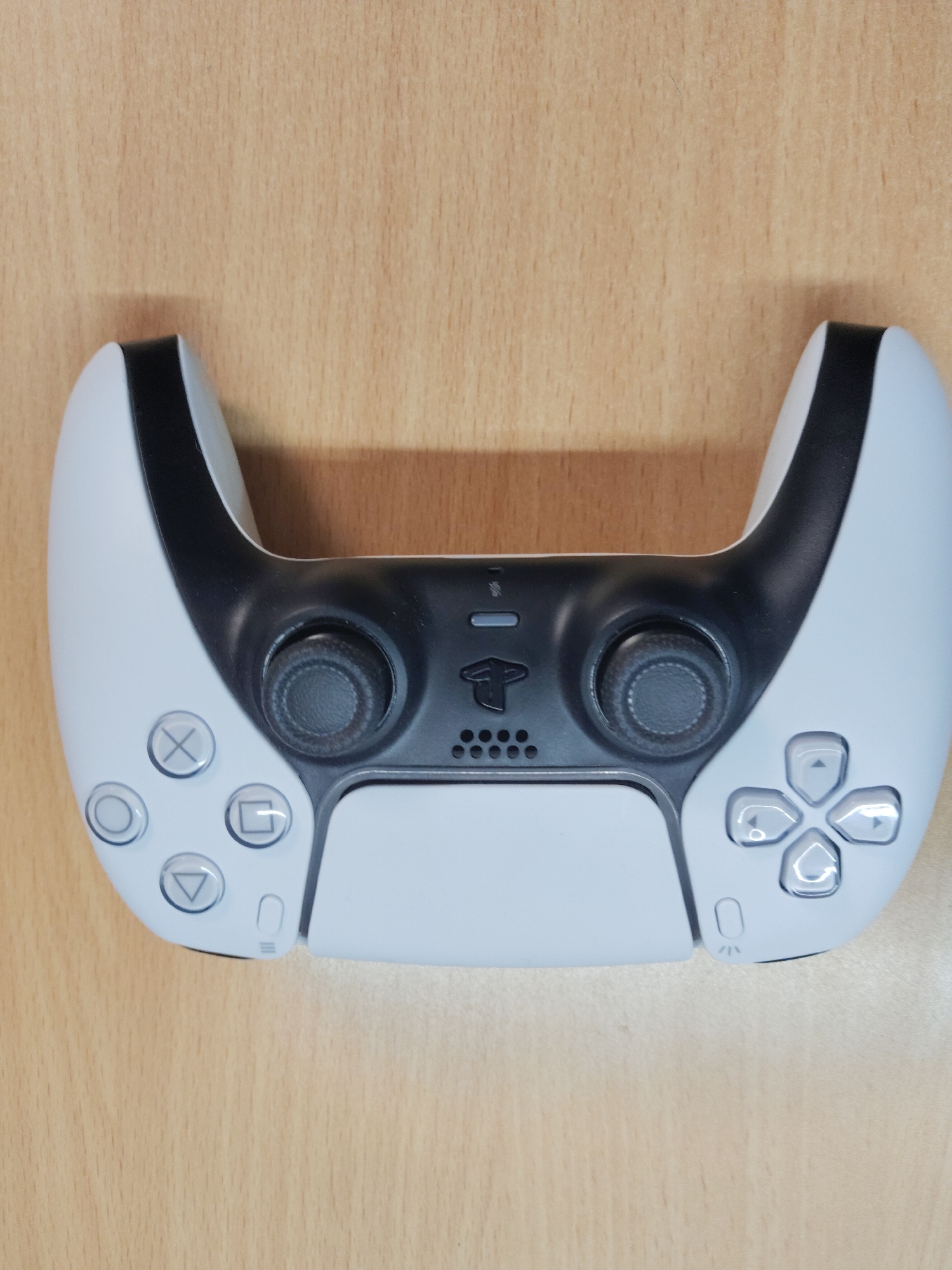 A white and black PlayStation 5 DualSense controller is placed on a light brown wooden surface. No location information is available.