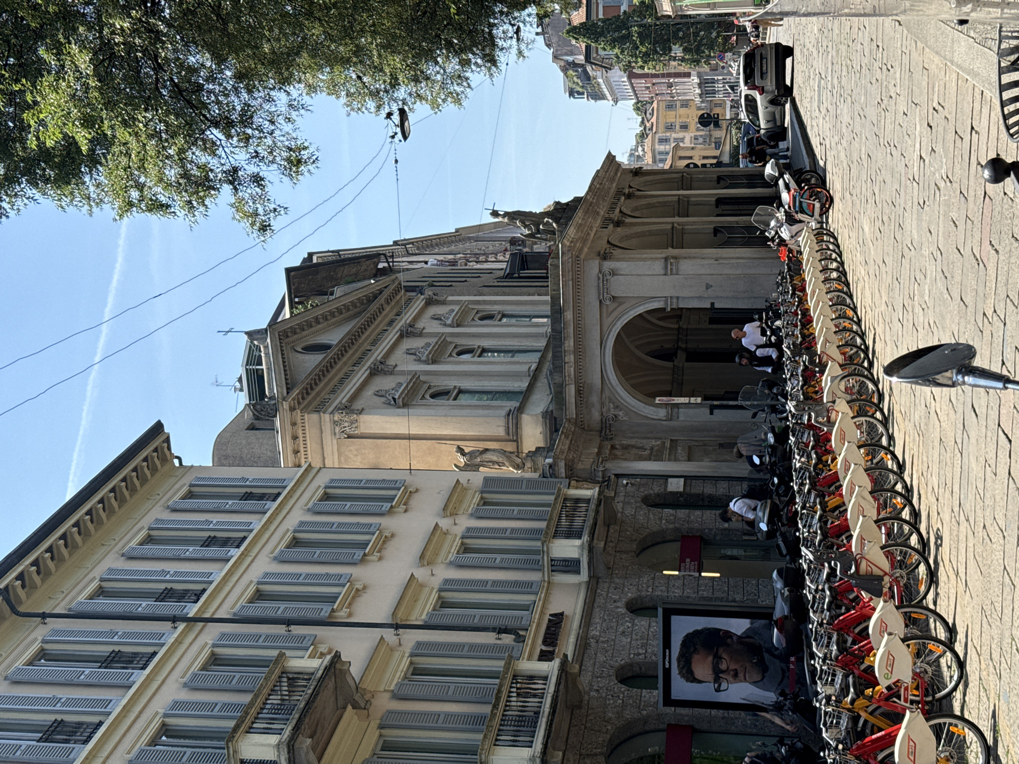 This is a medium shot, eye-level, horizontal photograph of a cobblestone street lined with red and yellow rental bicycles. On the left side of the street, a light-colored building with many windows covered by grey shutters is visible. To the right of this building, a stone facade with arched doorways and statues is present. Beyond this, a street continues with more buildings in the distance. The sky is clear blue with a few wispy clouds. Several people are visible in the scene, some standing and some seated on motorcycles. A car is parked further down the street. The overall impression is a sunny day in a European city.
