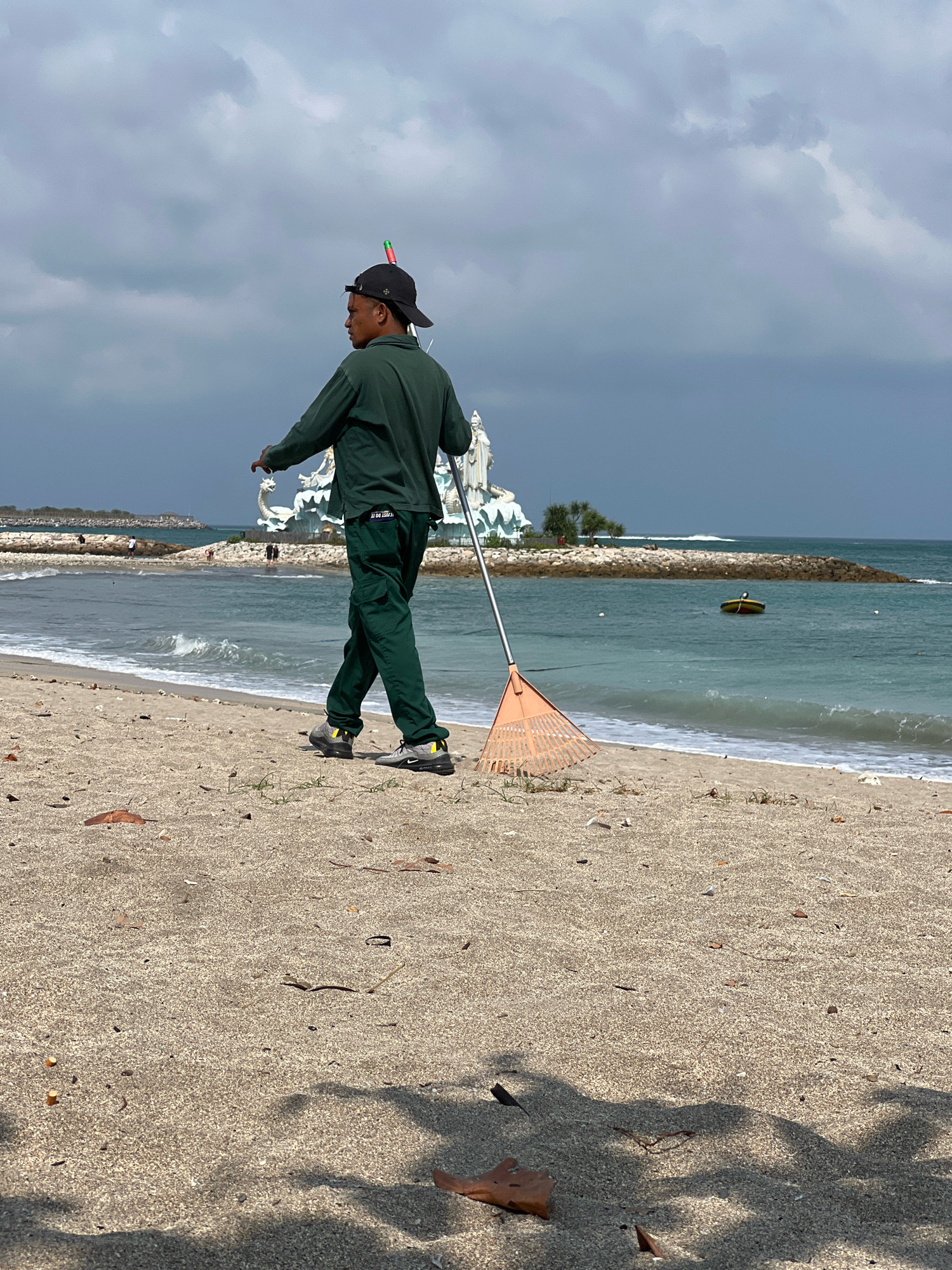 A man dressed in a dark green long-sleeved shirt and matching cargo pants, wearing a black baseball cap with a small colorful object protruding from it, walks along a sandy beach in Kuta, Indonesia. He is holding a long-handled orange rake with his right hand. The rake is positioned behind him and slightly to his right, with its tines resting on the sand. The man's left arm is bent at the elbow, and his hand is held in front of his body, appearing to be in motion. He is looking away from the camera, towards the ocean.
The beach is composed of light brown sand, with scattered debris such as leaves and small twigs. The ocean is a vibrant turquoise color, with gentle waves breaking near the shore. In the background, a man-made jetty or breakwater made of dark rocks extends into the sea. Beyond the breakwater, a white structure with elaborate sculptures, possibly a religious monument or a decorative landmark, is visible. Further out in the water, a small yellow and red boat is anchored. The sky above is filled with a mix of gray and white clouds, suggesting an overcast or partially cloudy day. The overall scene depicts a man engaged in cleaning the beach on a cloudy day in Kuta.