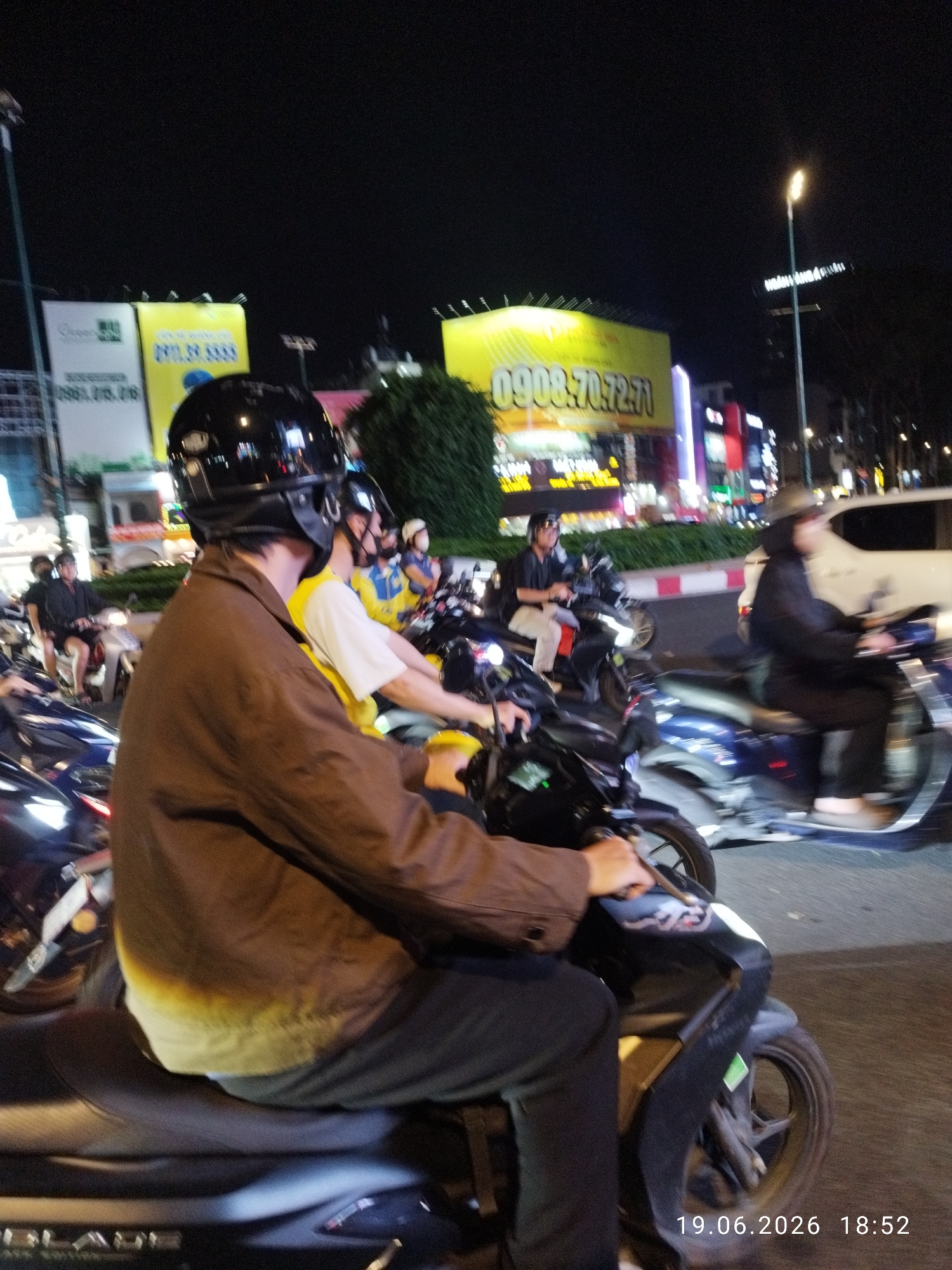 The image depicts a nighttime street scene in Thủ Đức City, Vietnam, likely during rush hour or a busy period. The foreground is dominated by a person riding a black motorcycle, wearing a black helmet and a brown jacket. The motorcycle has a dark frame and a portion of the wheel is visible. Behind this rider, another person on a motorcycle is visible, wearing a yellow shirt and a black helmet. Further back, several other motorcycles and riders are present, some with passengers. The background features illuminated billboards and storefronts, indicative of a commercial area. One prominent billboard displays text in Vietnamese and a phone number: "0908:70.72.71". Another billboard on the left shows a green logo and text that includes "0961.015.06". A street light illuminates the scene from the right. The overall impression is one of a bustling urban environment at night, with a significant number of motorcycles on the road. The date and time stamp "19.06.2026 18:52" is present in the bottom right corner.