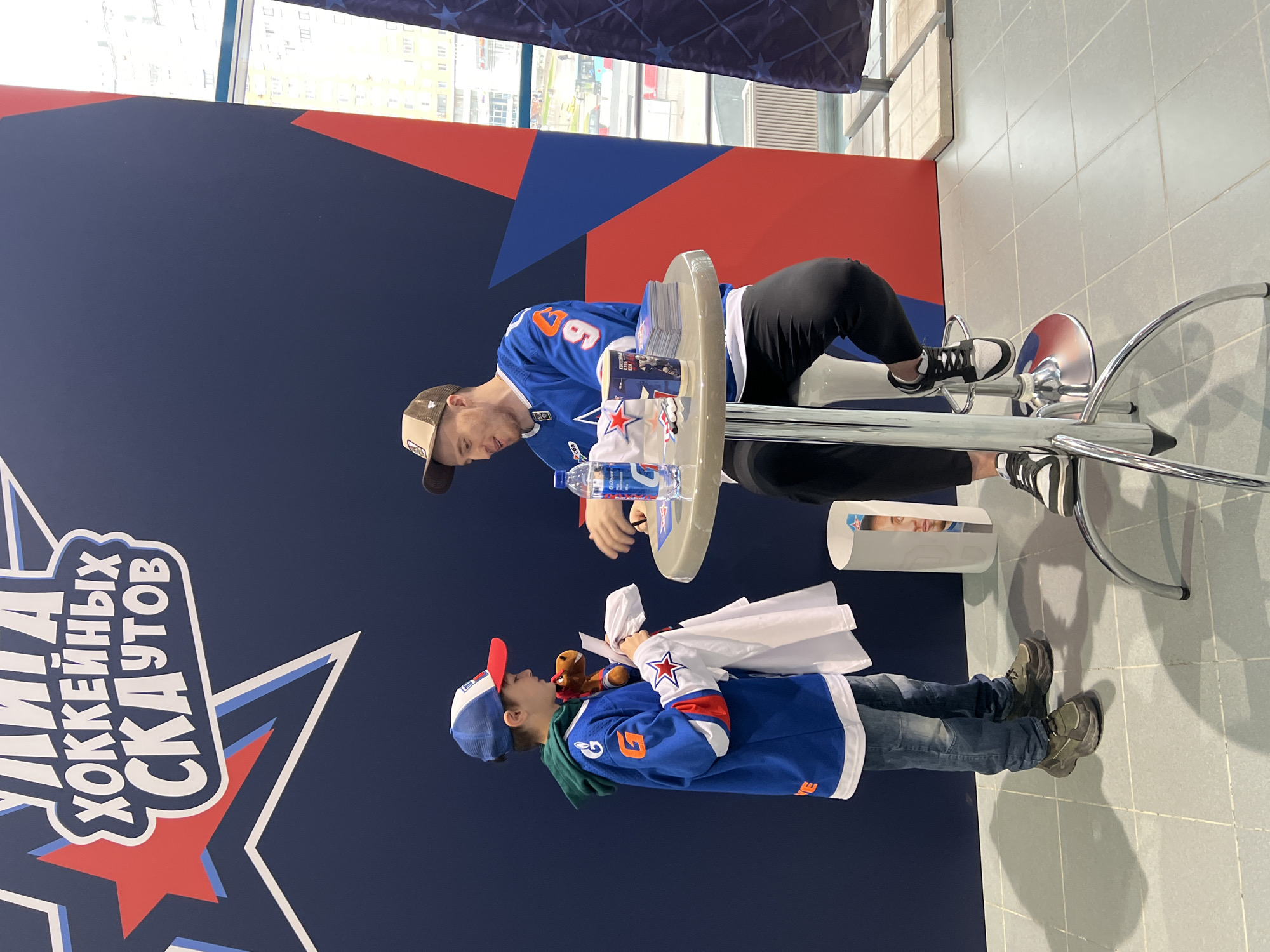 The image captures a scene involving a hockey player signing an autograph for a young fan. The setting appears to be indoors, likely at a promotional event or fan meet-and-greet.
The hockey player, wearing a blue jersey with the number 67, is seated on a bar-style stool at a small table. He is focused on signing an item for the young fan. On the table are several signed cards, a water bottle, and a white towel with a star emblem.
The young fan, also wearing a blue hockey jersey, is holding a small stuffed animal and another white towel with a star emblem.
The background features a large dark blue wall adorned with the logo for "SKA Hockey Club" or "Saint Petersburg Army Club," a professional ice hockey team based in Saint Petersburg, Russia. The wall also has a red and blue design element.
The floor is tiled in light grey. There is a window in the background. Overall, the image captures a positive interaction between a sports figure and a young supporter.